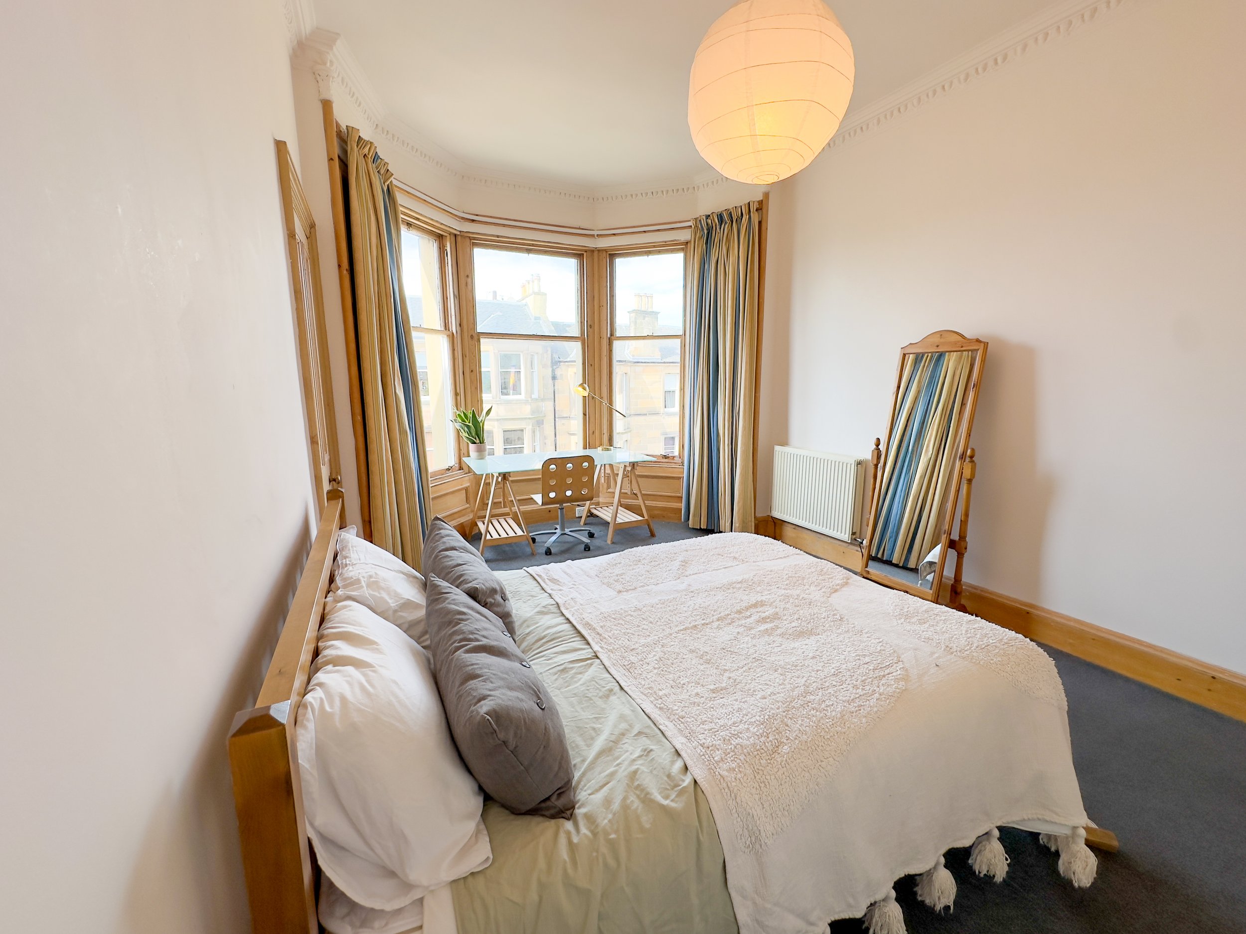A bedroom with a wooden bed, beige bedding, and throw pillows. A large window with striped curtains, a small desk with a potted plant, and a standing mirror leaning against the wall.