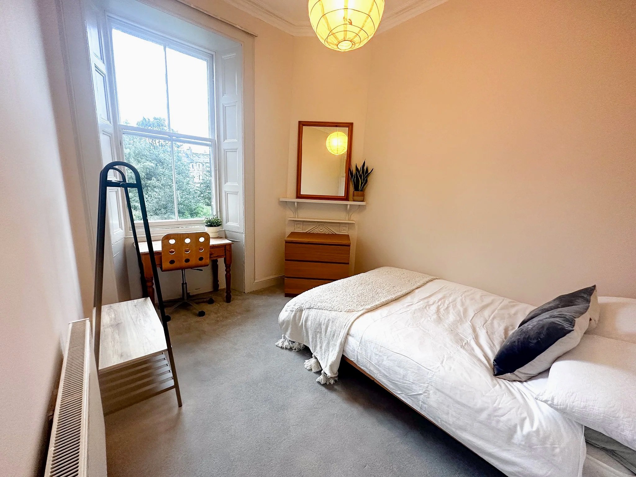 A small bedroom with a bed, a dresser, a mirror, a window with a view of trees, and a desk by the window. The room has a yellow paper lantern ceiling light and neutral-colored walls.