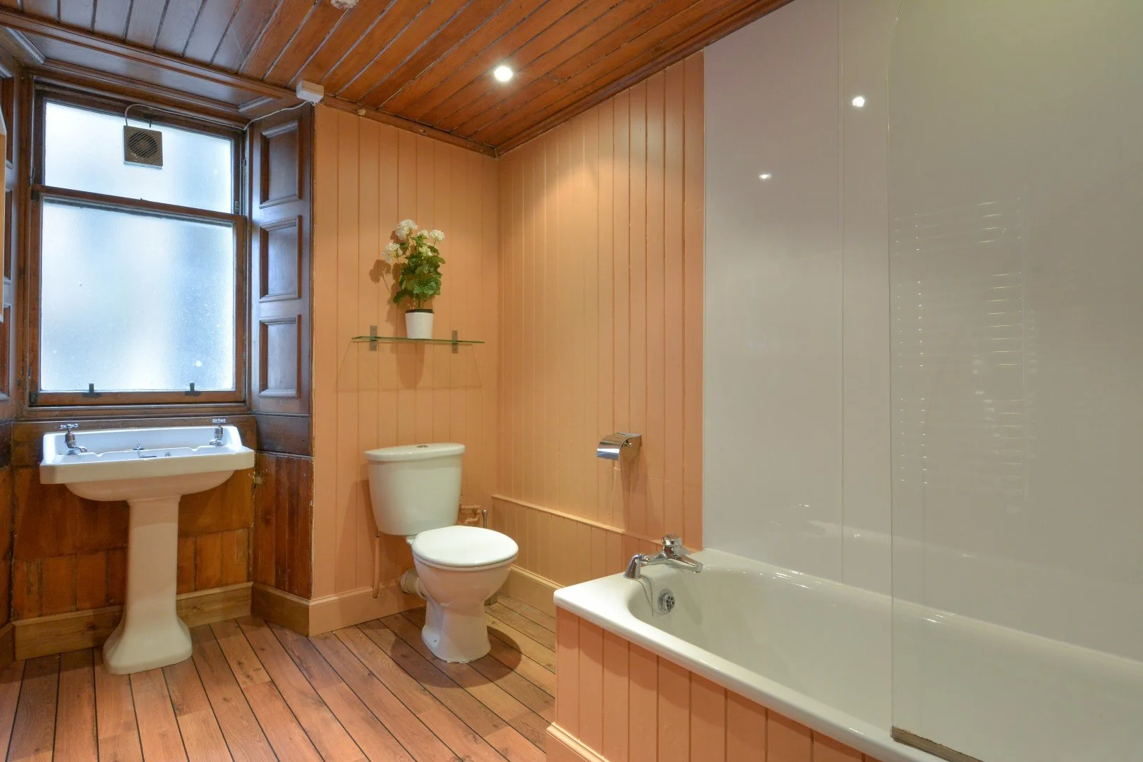 Bathroom with wood-paneled walls and floors, featuring a frosted window, a pedestal sink, a toilet, a bathtub with a glass shower screen, a wall-mounted shelf with a potted plant, and recessed ceiling lights.