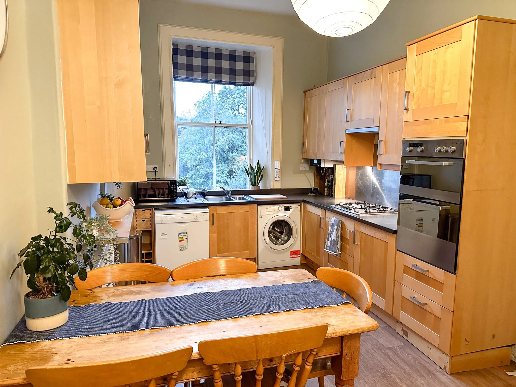 A cozy kitchen with wooden cabinets, a wooden dining table with chairs, a window with a blue checkered curtain, and various kitchen appliances including a washing machine, oven, microwave, toaster, and a bowl of fruit.