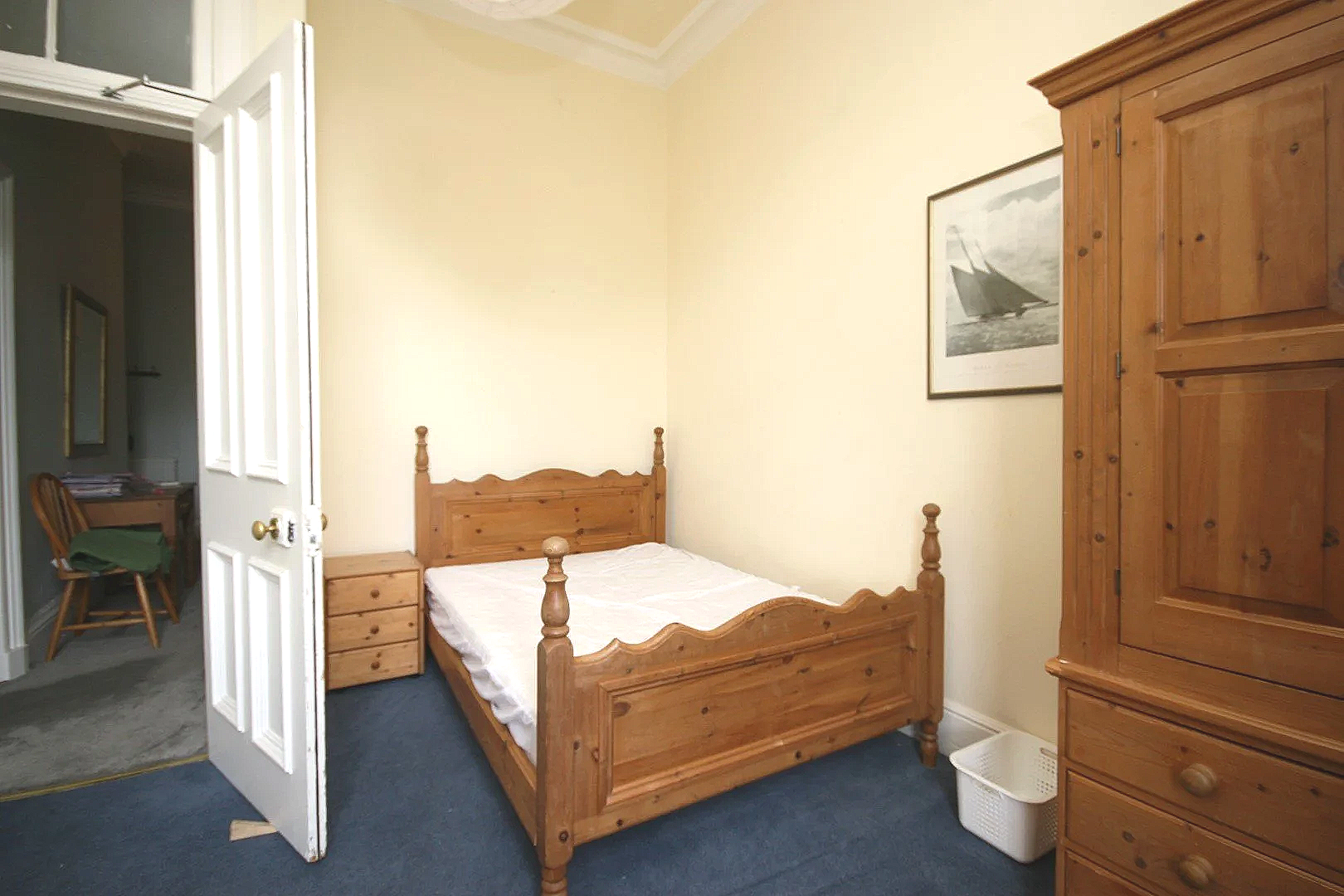 A bedroom with a wooden bed frame, a small wooden nightstand, a wardrobe, a framed picture of a sailboat on the wall, and a white laundry basket on the floor.