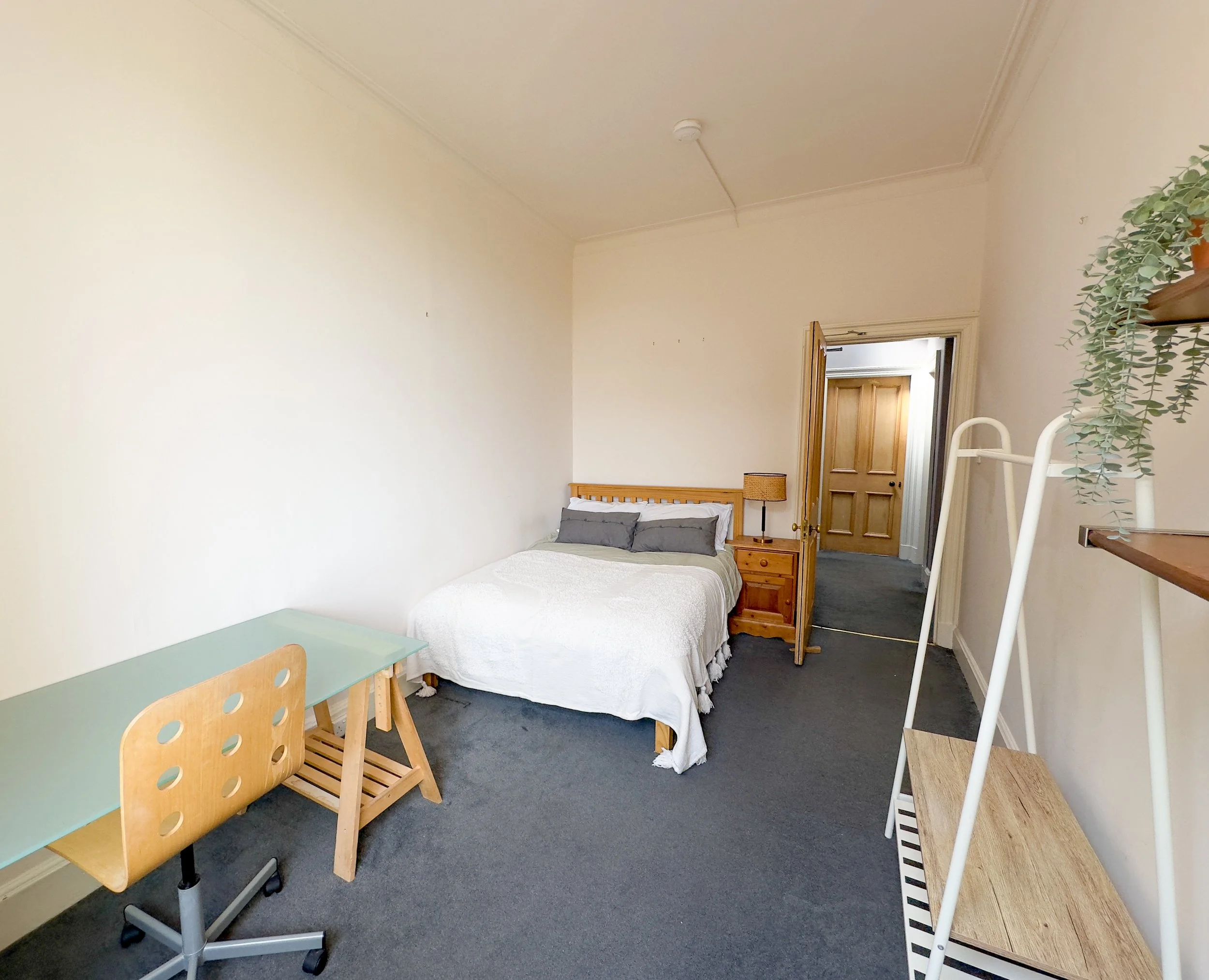 A bedroom with a bed, nightstand, and desk, featuring neutral tones and minimal decor.