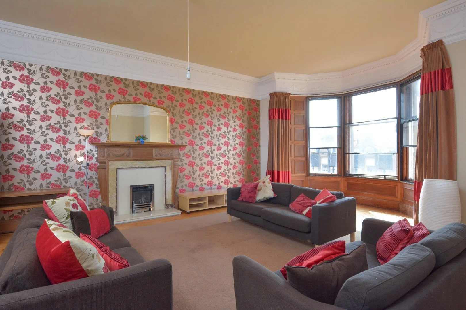 Living room with floral wallpaper, wooden fireplace mantel with mirror, large bay window with curtains, three sofas with red and patterned pillows, beige carpet, and a white floor lamp.