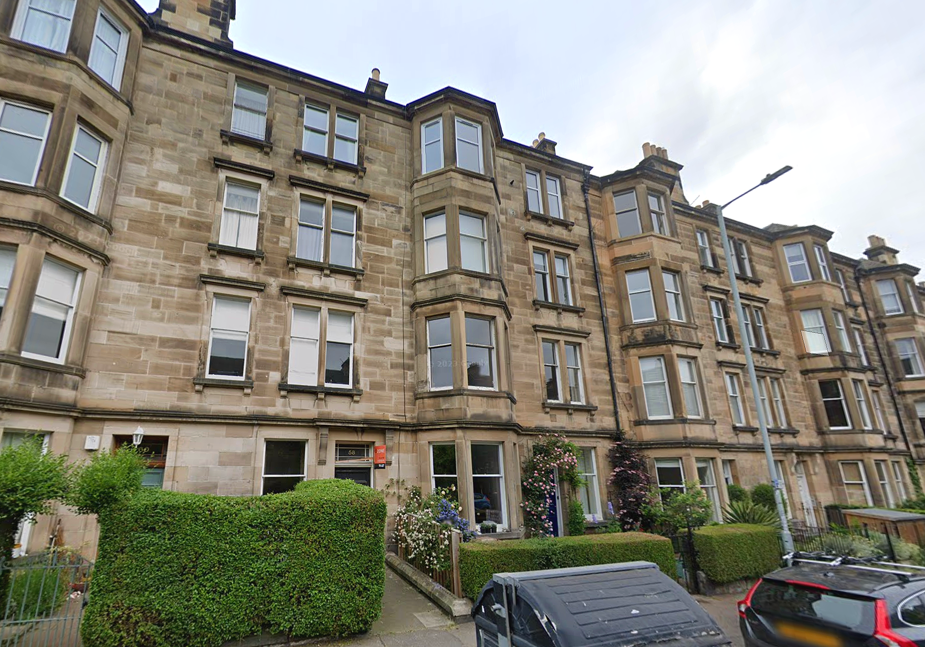 View of a multi-story apartment building made of sandstone with bay windows, garden areas, and parked cars in front.