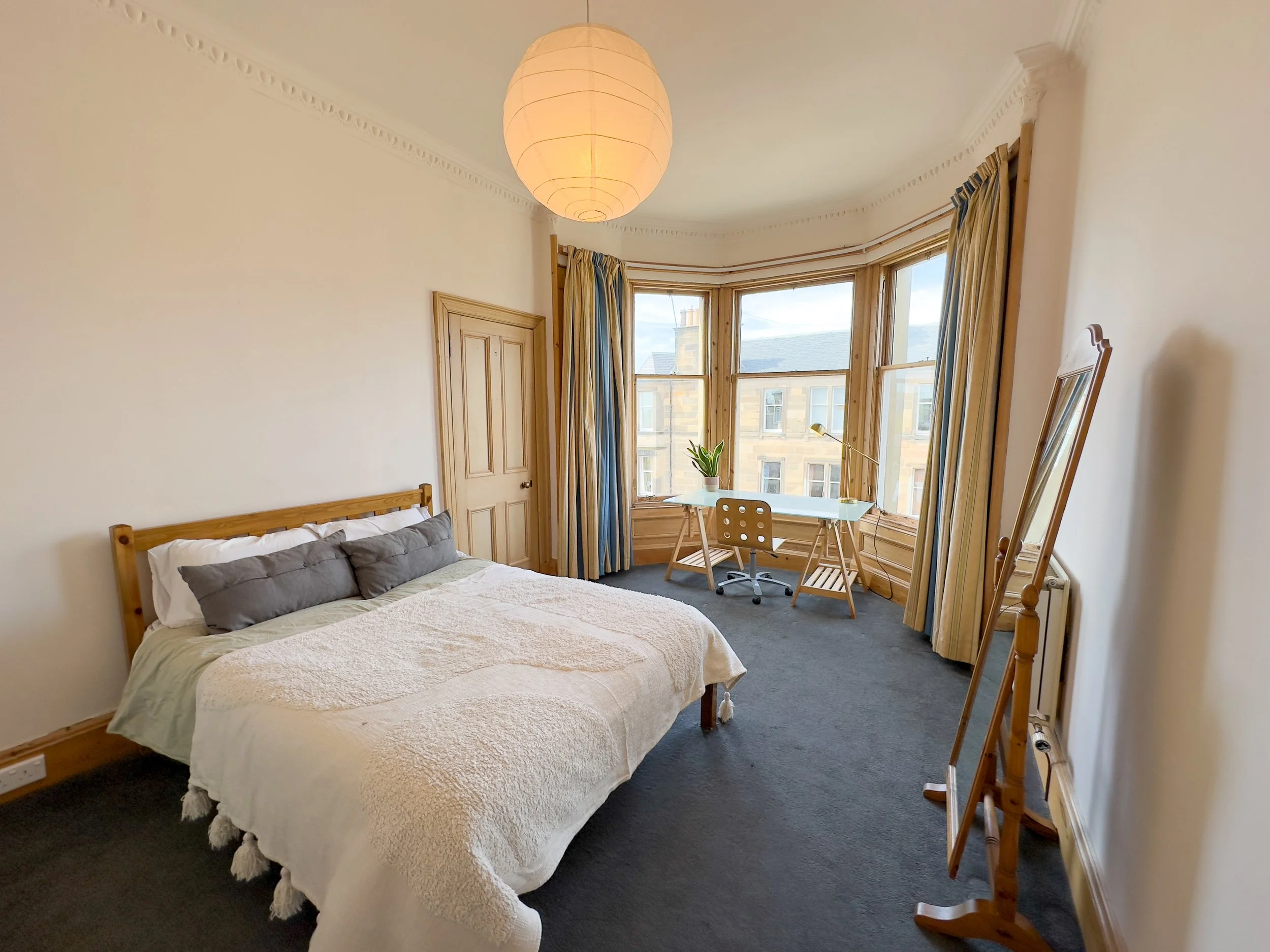 A bedroom with a bed, wooden headboard, and gray pillows. A desk with a plant and a lamp near the large bay window. A mirror on an easel, curtains, and a paper lantern ceiling light.