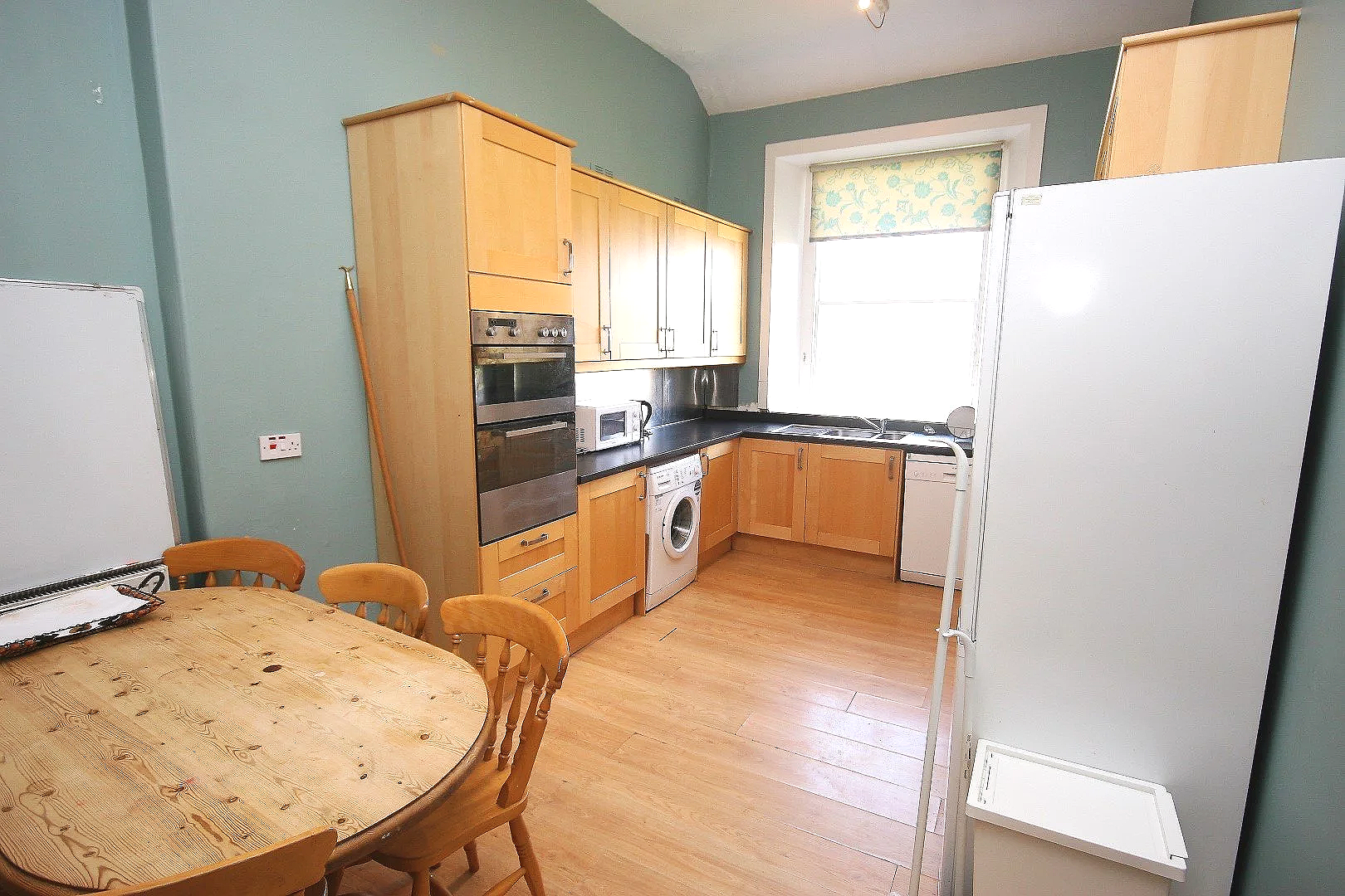 Kitchen with wooden cabinets, black countertops, a window with floral blind, and light-colored flooring. Includes a dining table with four wooden chairs and appliances like a microwave, washing machine, and refrigerator.