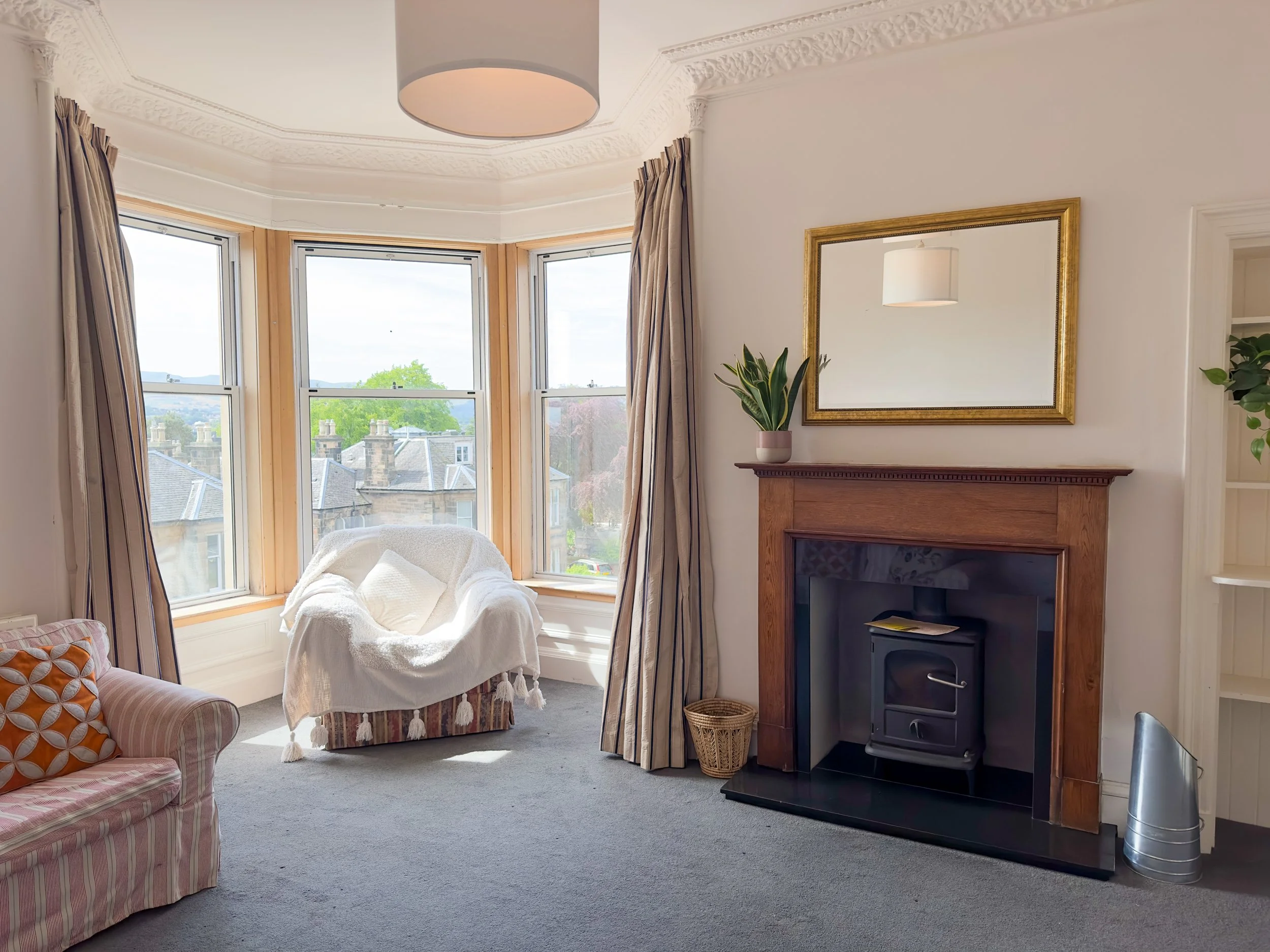 A living room with a large bay window, cream and beige curtains, a plush armchair with a throw blanket, a patterned sofa with a pillow, a mirror above a fireplace, a potted plant, and a wood-burning stove.