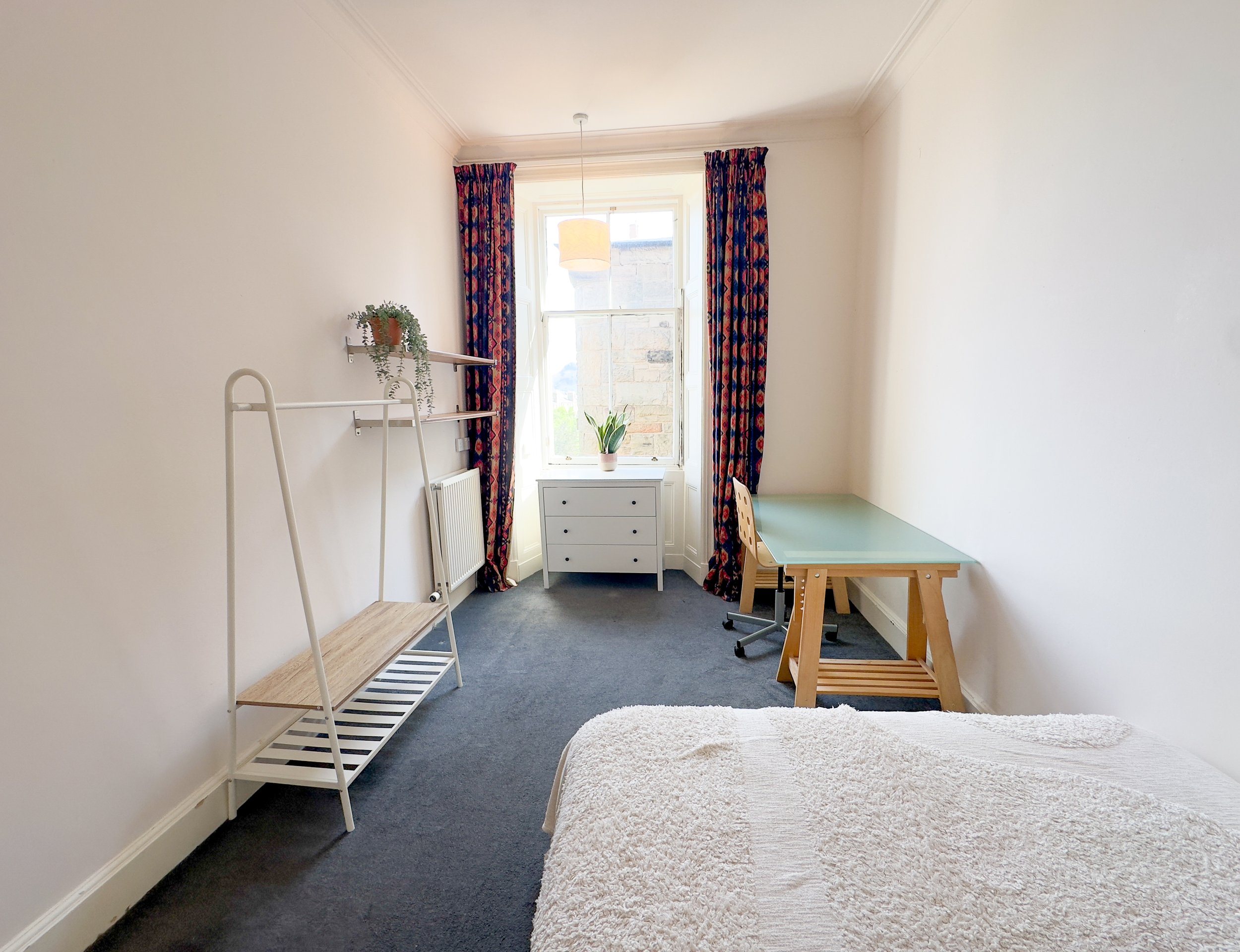 A bedroom with white walls, dark carpet, and a window with colorful patterned curtains. The room has a white bed with a beige blanket, a green desk with a wooden chair, a white chest of drawers, and a metal shelf with a potted plant. There is a hanging light fixture and a radiator near the window.