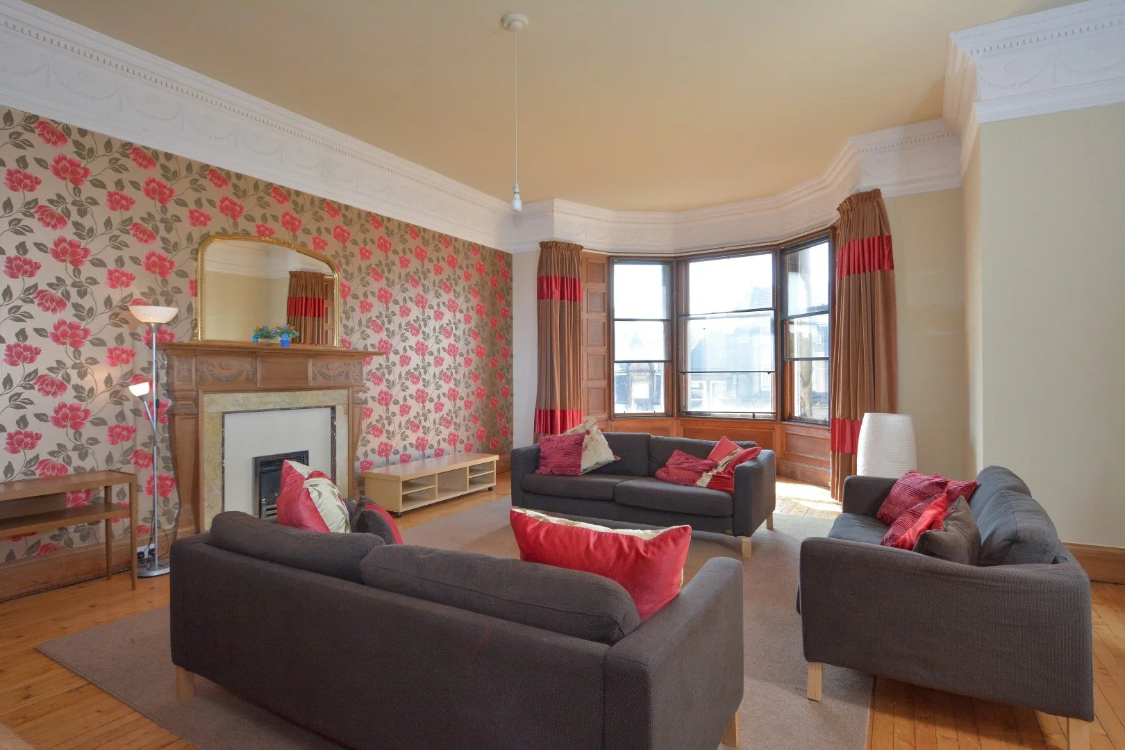 Living room with floral wallpaper, three dark sofas with red and cream cushions, wooden flooring, fireplace with a mirror above, large bay window with red and brown curtains, and a tall floor lamp.