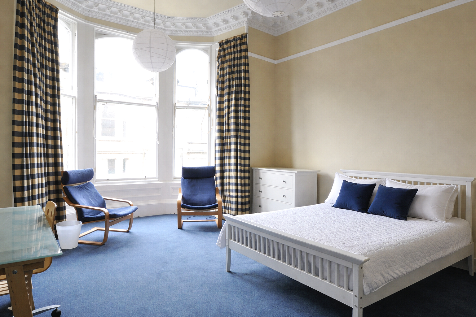 A bedroom with a white bed, navy pillows, a white dresser, and navy chairs. Large bay window with checkered curtains, blue carpet, and decorative ceiling molding.