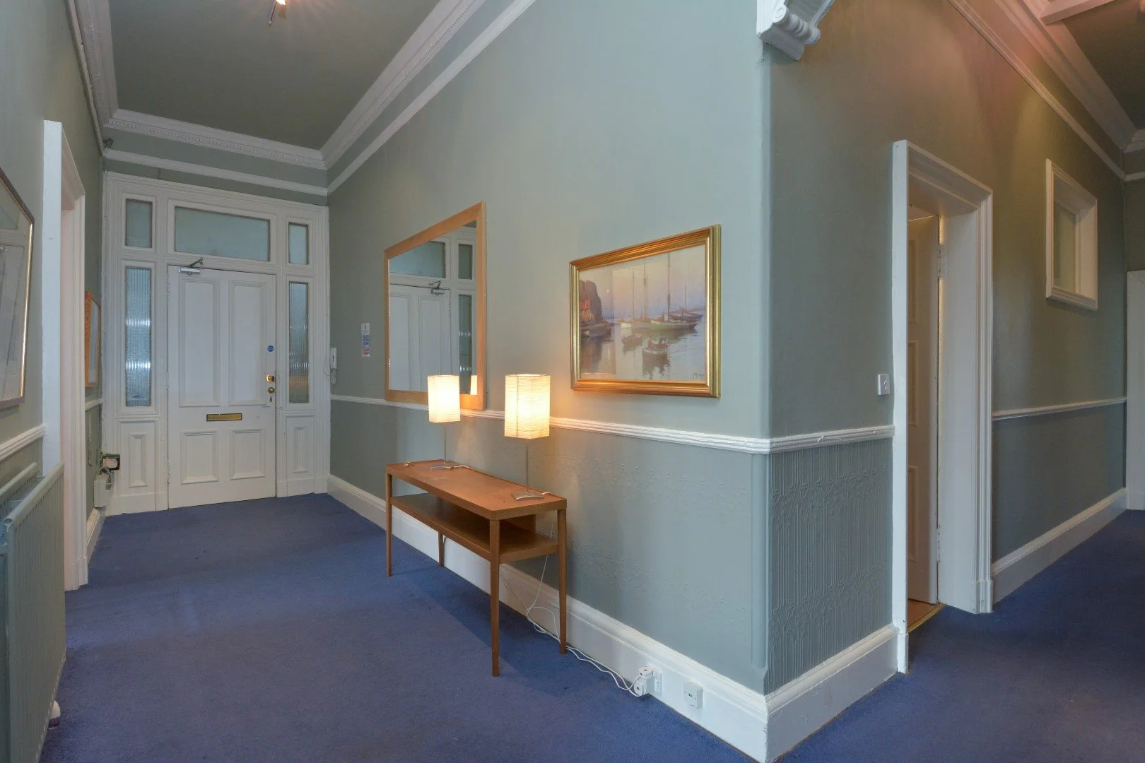 Interior of a house entryway with front door, blue carpet, a wooden table with two lamps, framed artwork, and hallways leading to other rooms.