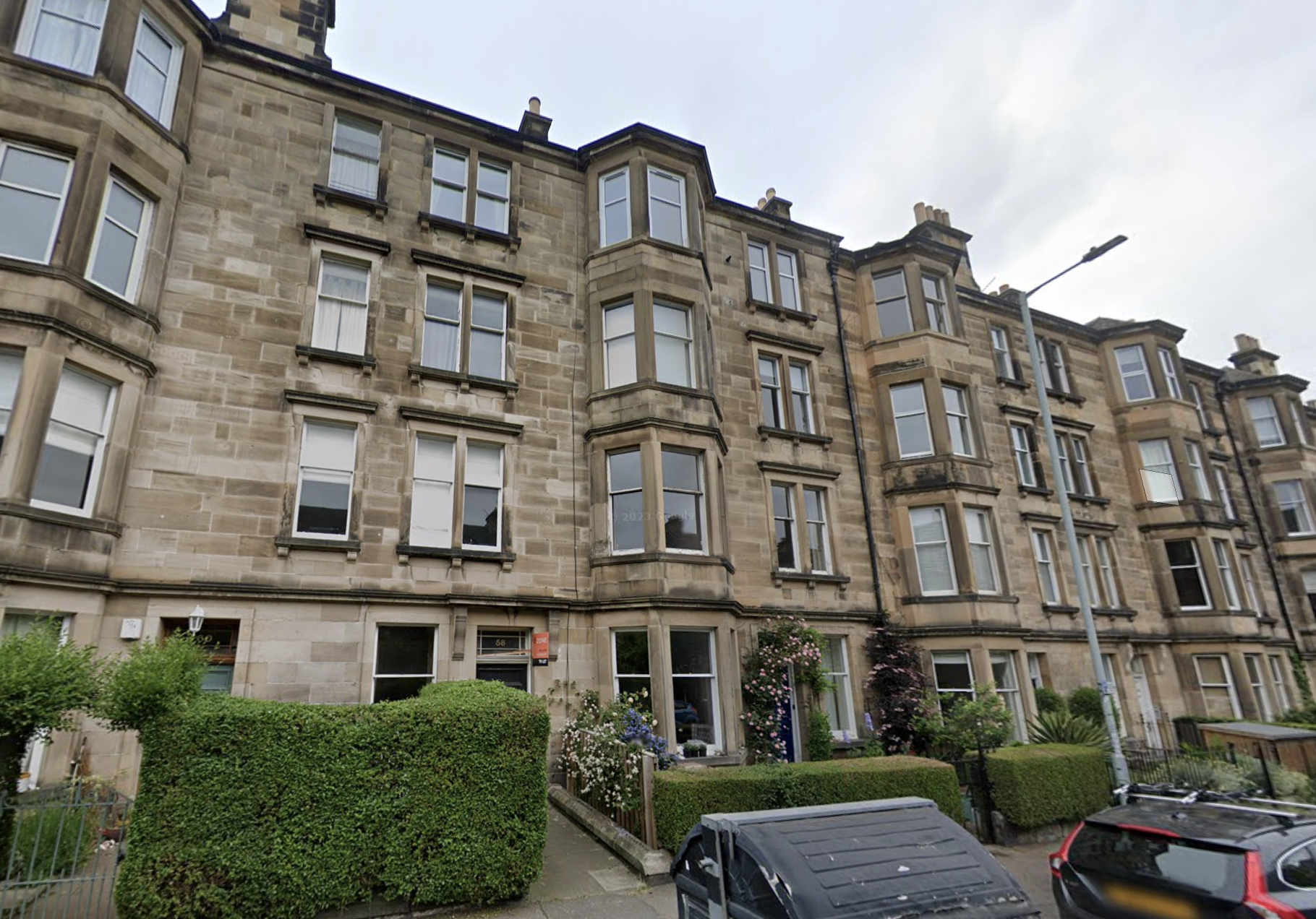 Four-story brownstone apartment building with bay windows, surrounded by greenery and parked cars