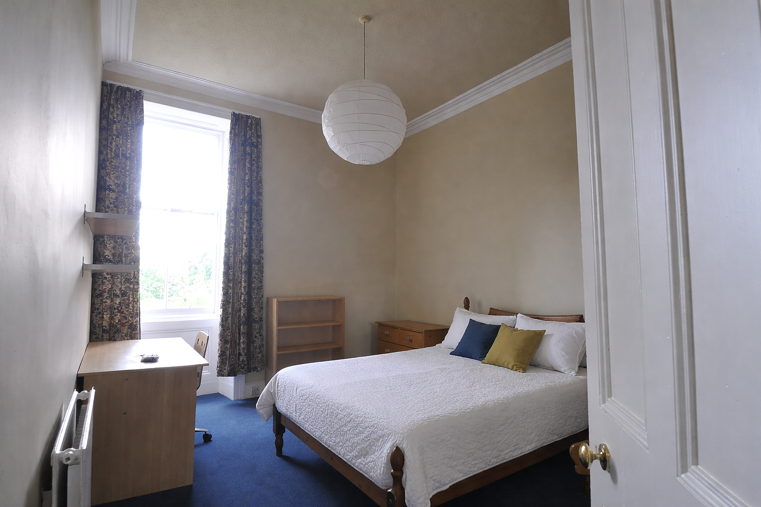 A bedroom with a bed, pillows, window with curtains, a small desk, a chair, a bookshelf, and a dresser, with white walls and blue carpet.