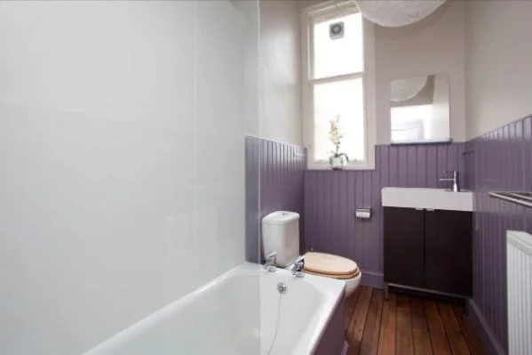 A small bathroom with a bathtub, toilet, and a vanity with a sink, purple wainscoting, a window with a plant, and hardwood flooring.