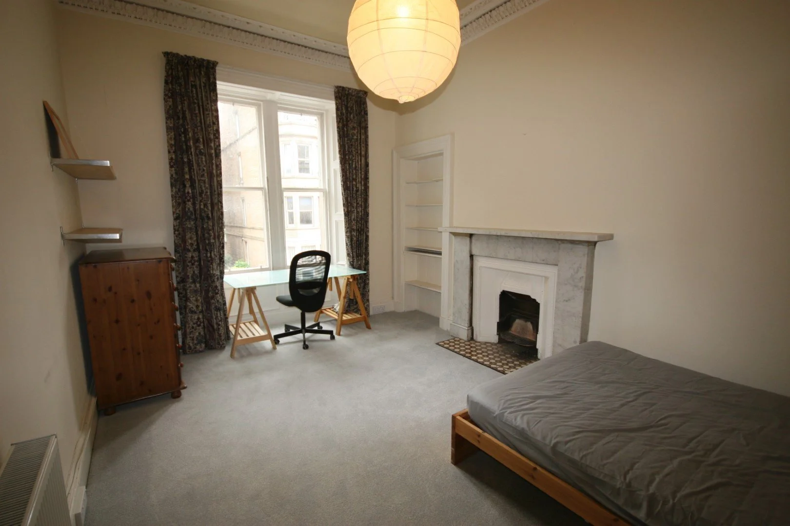 Modern wooden house on a grassy landscape with small trees near a lake, with mountains in the background under a cloudy sky.