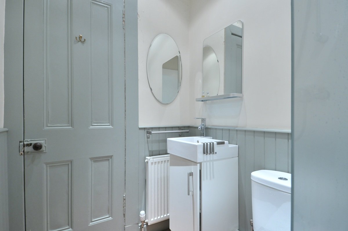 Small bathroom with a light gray door, a white sink with a cabinet, a mirror, a small wall-mounted shelf, a white toilet, and a radiator.