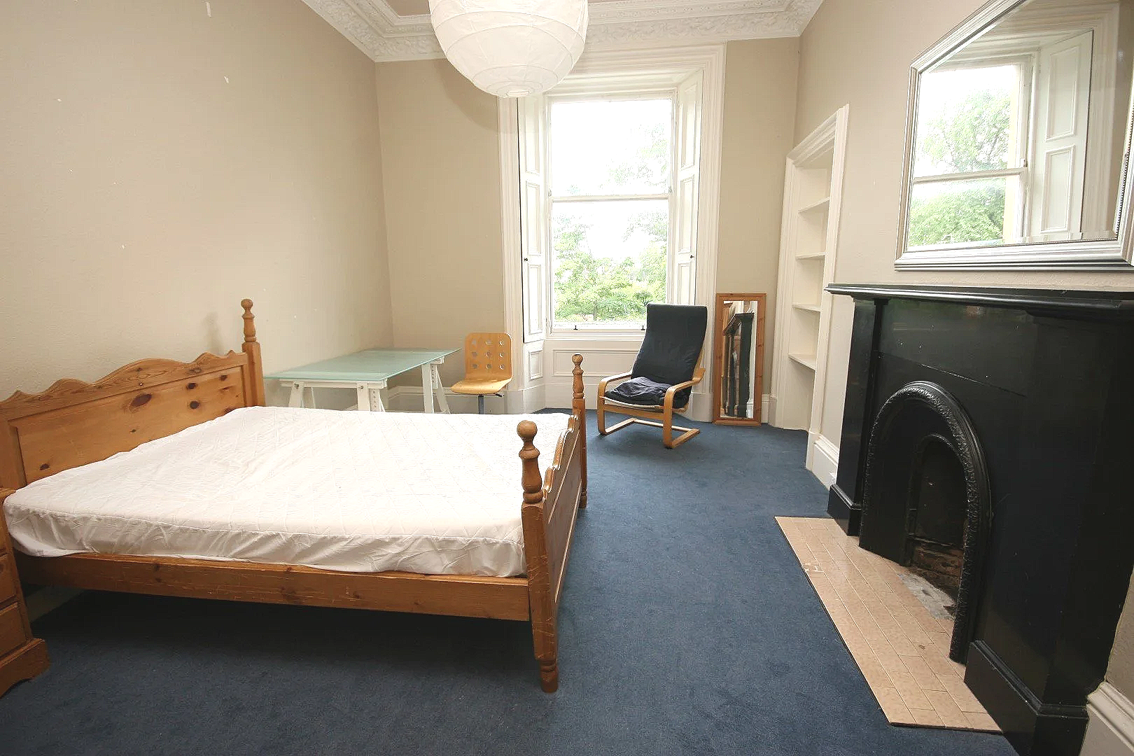 A bedroom with a wooden bed and a white mattress, a small glass table, two chairs, a black armchair, a wooden framed mirror, a white bookshelf, a window with shutters, a black fireplace, and a blue carpet.