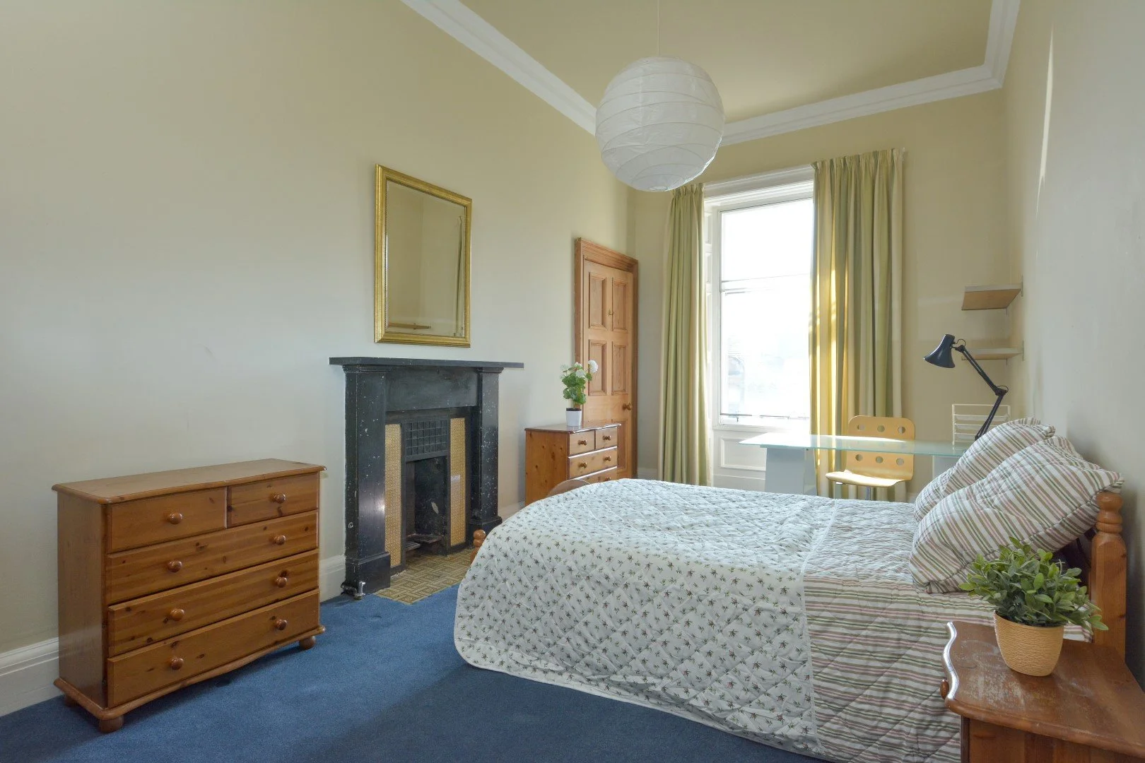 A bedroom with a bed, wooden furniture, a black fireplace, a mirror, a window with yellow curtains, a desk with a lamp and chair, and a ceiling light