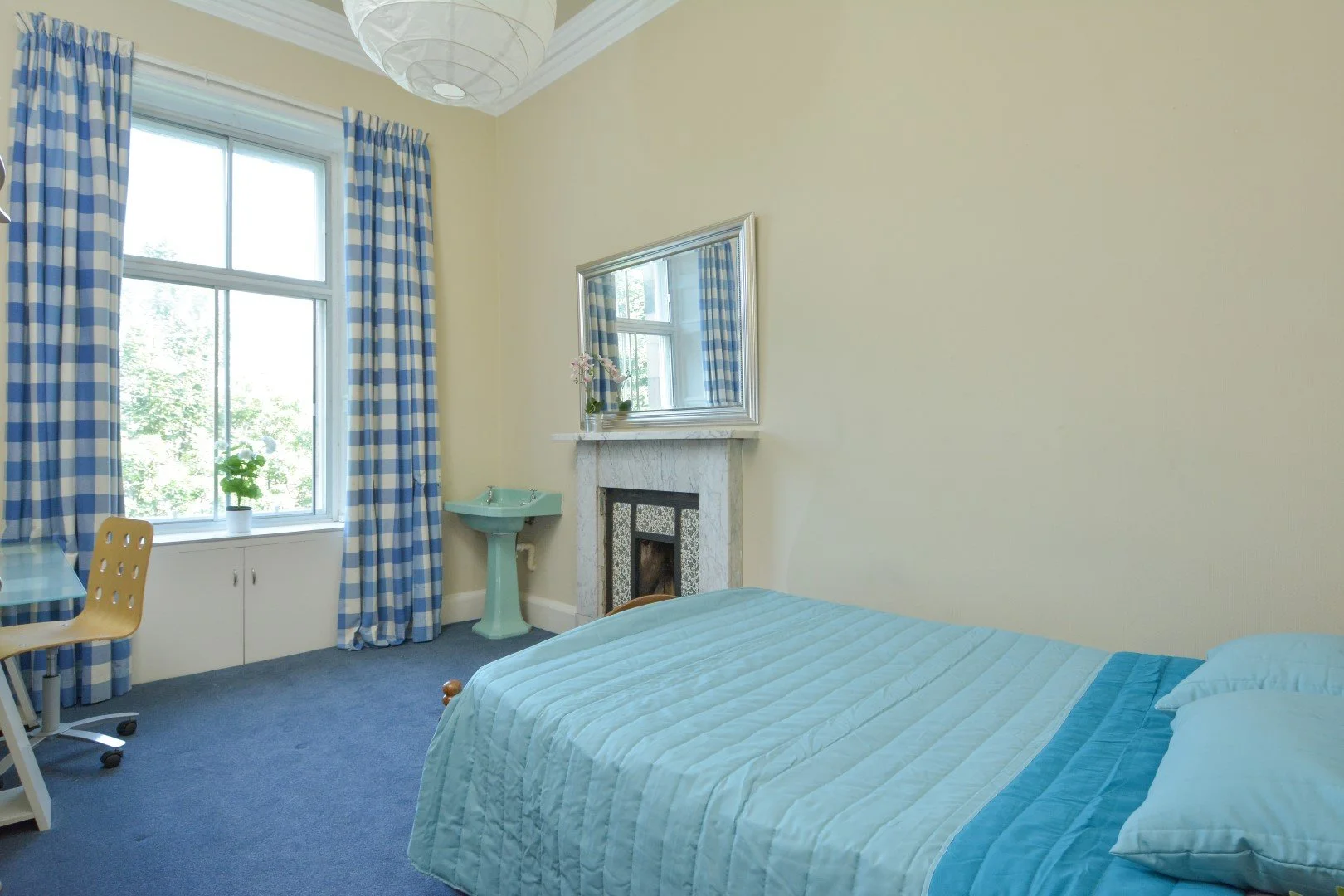 A bedroom with a blue bedspread, a large window with checkered blue and white curtains, a small table with a plant, a green sink, a fireplace with a large mirror above it, and a chair.
