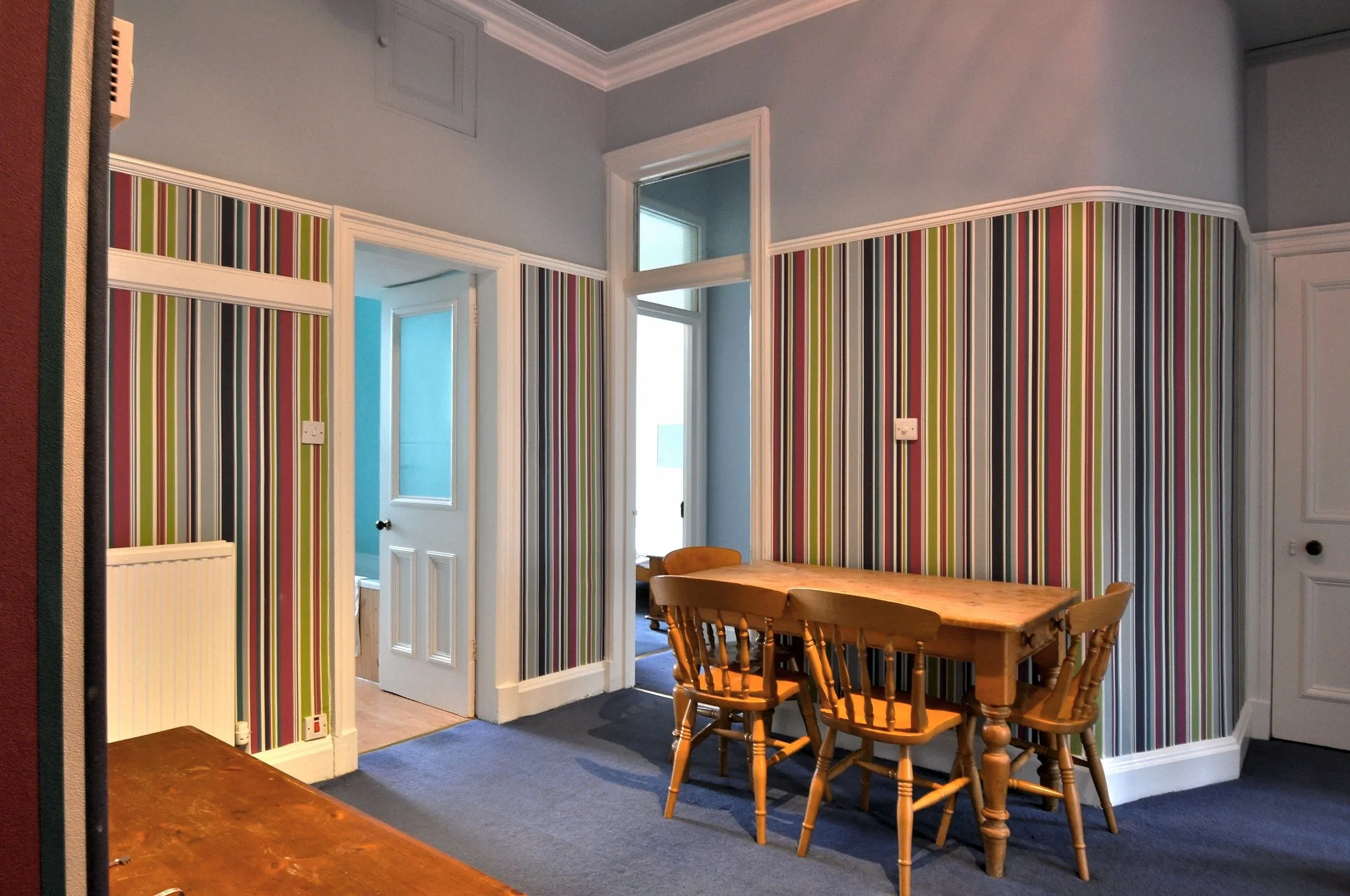 A dining area with a wooden table and four matching wooden chairs against a wall covered with colorful striped wallpaper. There are two doorways, one leading to a room with a window, and the other to a room with a closed door. The walls are painted a light blue, and the floor has blue carpeting.