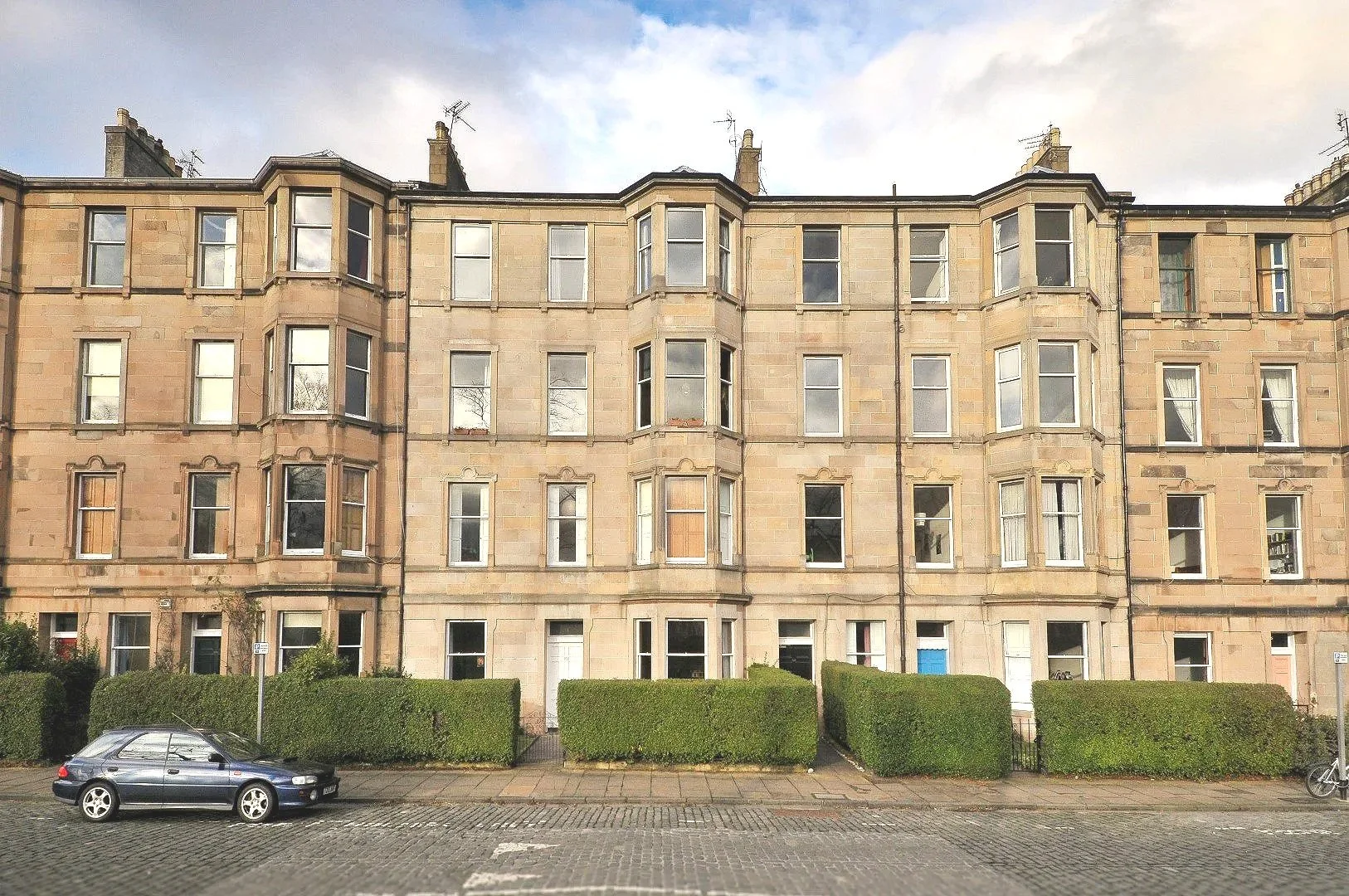 Living room with light green walls, a blue sofa with patterned and solid pillows, a wooden dining table with six chairs, a wooden cabinet, a clock and some appliances near a large window.