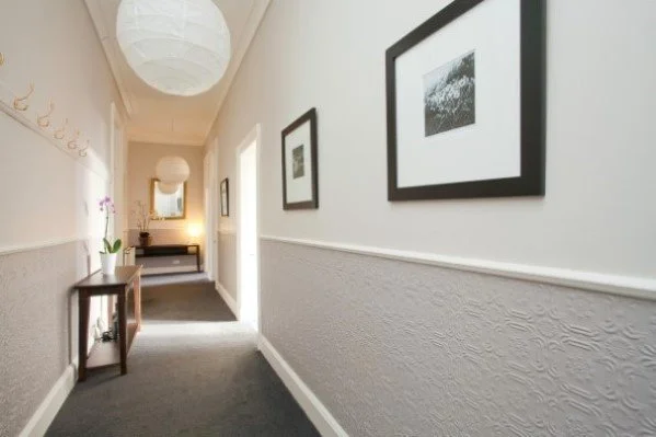 Hallway with gray carpet, black framed photos on the wall, a console table with a potted orchid, and a doorway leading to another room visible in the background.