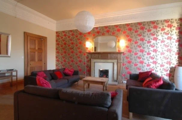 Living room with floral wallpaper, four dark sofas with red cushions, a wooden coffee table, fireplace, mirror above the fireplace, two wall-mounted lights, a hanging paper lantern light, wooden floor, and door on the left side.