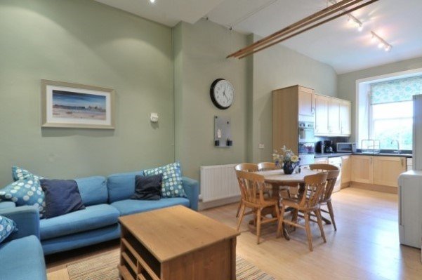 Living room with light green walls, a blue sofa with patterned and solid pillows, a wooden dining table with six chairs, a wooden cabinet, a clock and some appliances near a large window.