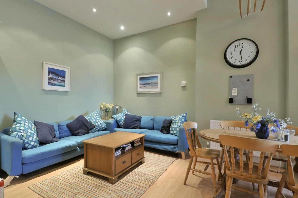 Living room with a light green wall, two framed landscape paintings, a blue sectional sofa with patterned and solid pillows, a wooden coffee table, a round wooden dining table with chairs, a vase of flowers, a wall clock, and a small bulletin board.