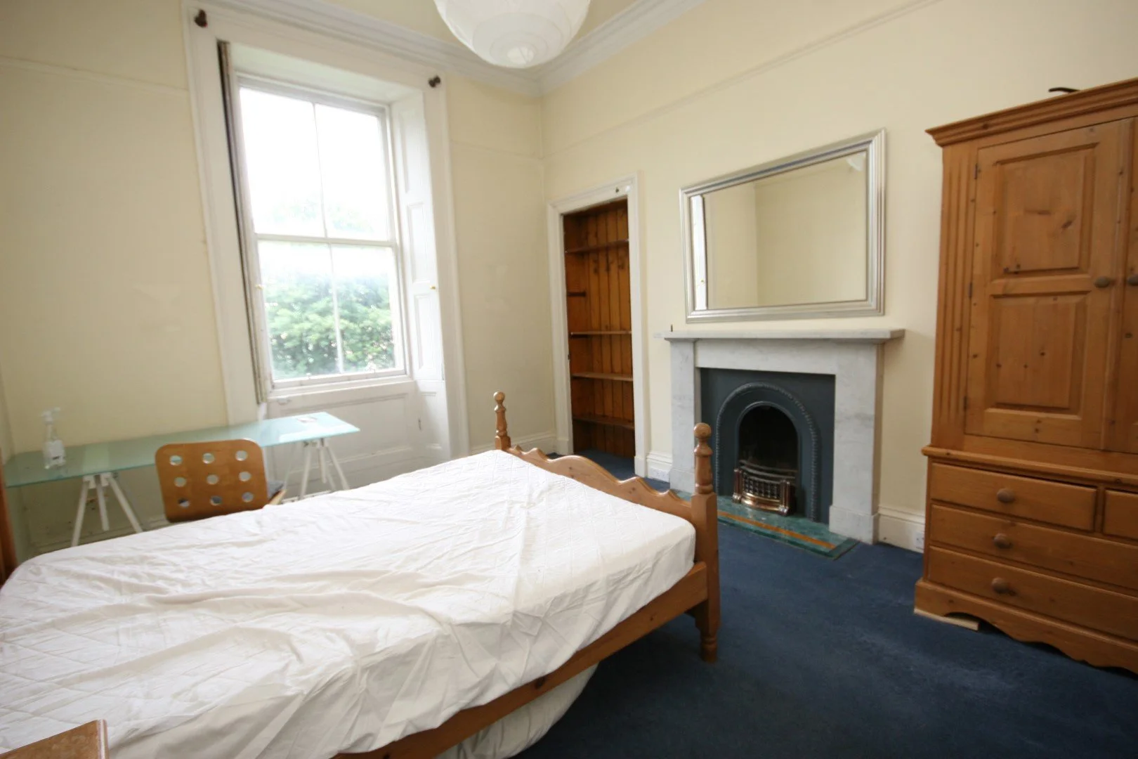 Bedroom with a single bed, wooden wardrobe, fireplace with a mirror above, window with white shutters, small glass table with a bottle, and a wooden chair with a cutout pattern.