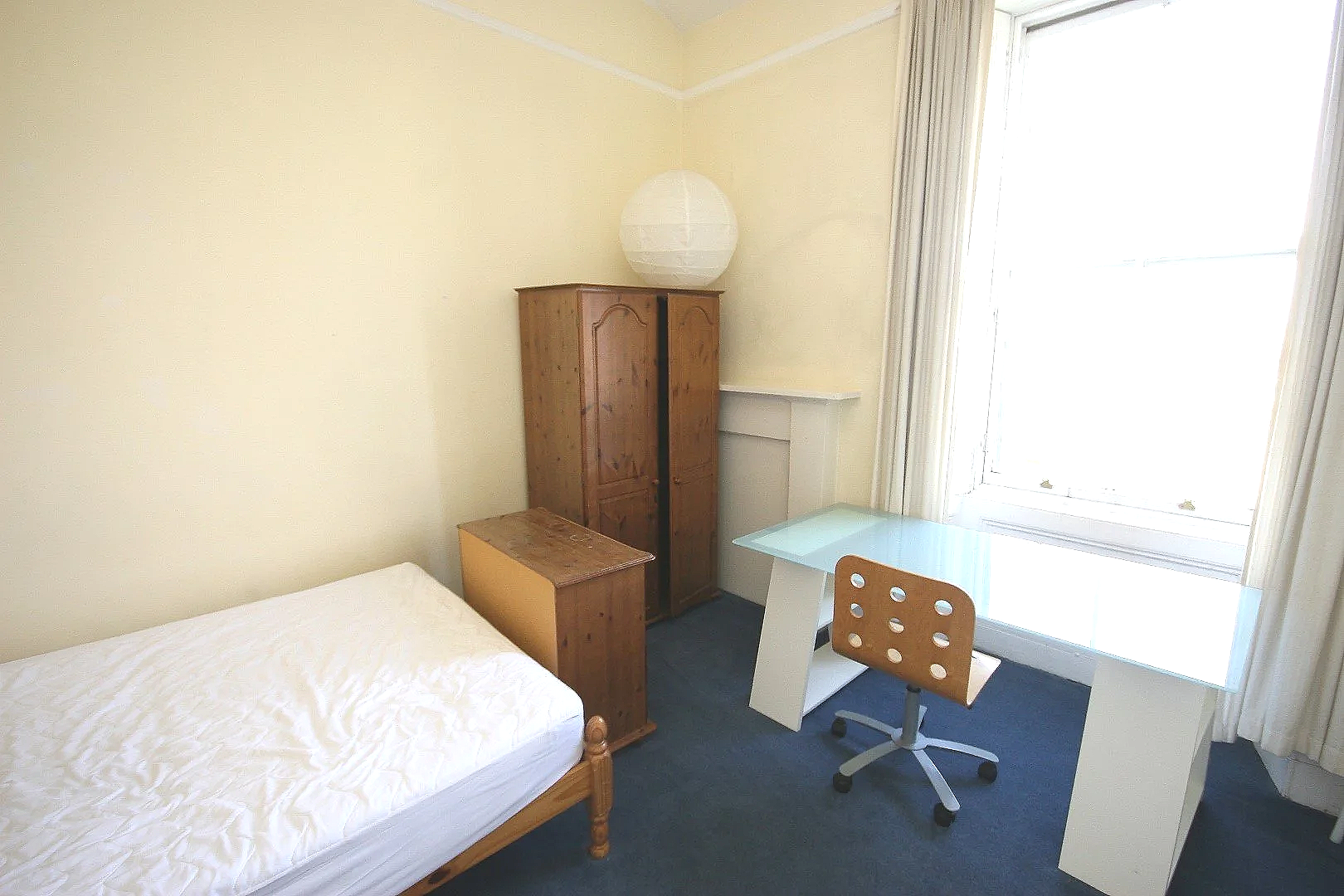 A simple bedroom with a wooden bed frame and mattress, a small wooden cabinet, a wardrobe, a desk with a glass top, an office chair, and a large window with curtains.