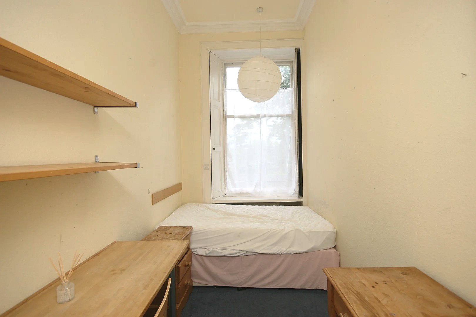 Small bedroom with a single bed, a window with sheer curtains, and wooden furniture including a desk and shelves.
