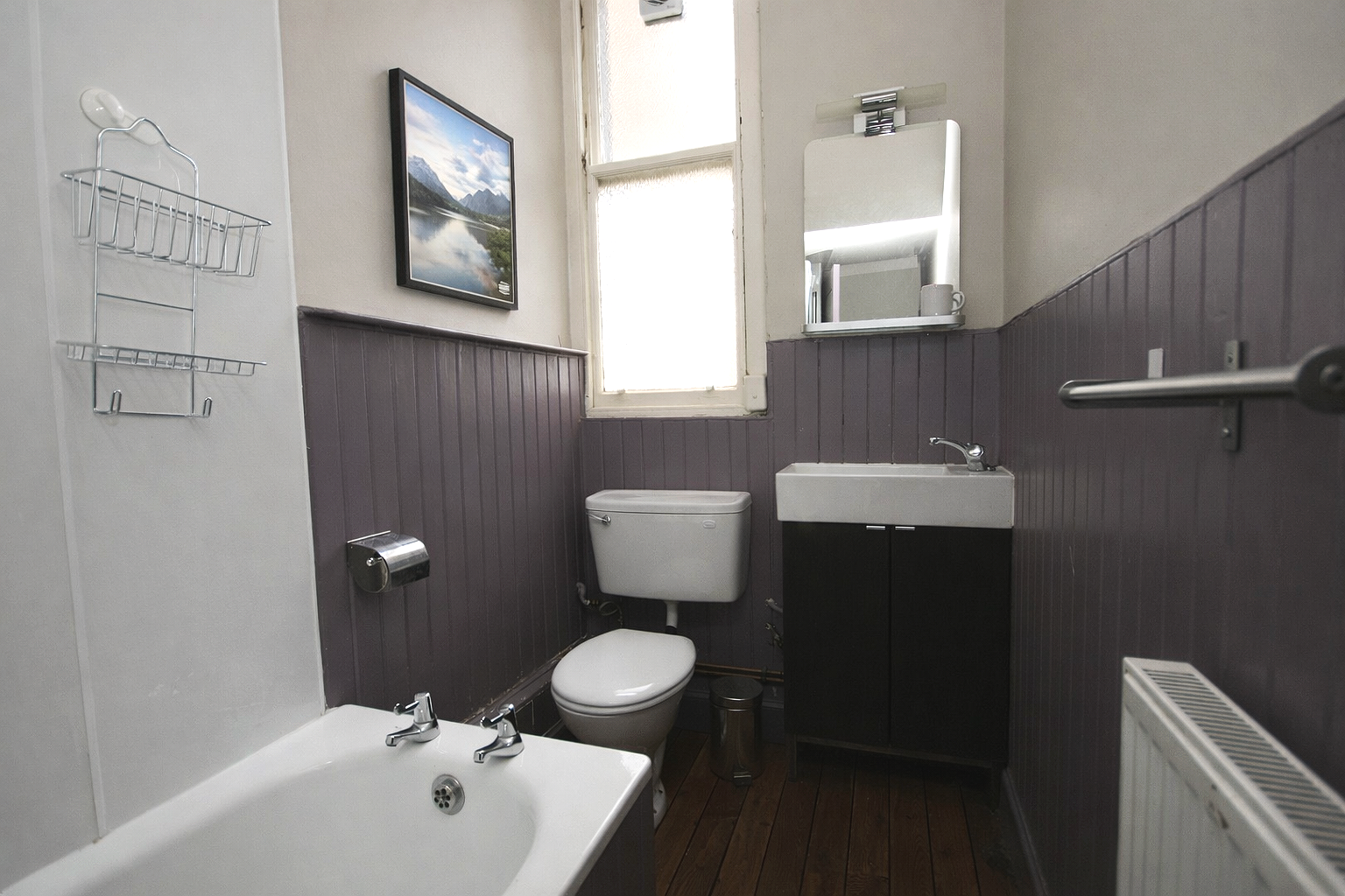Small bathroom with purple wainscoting, a toilet, a white sink with black cabinet, a mirror, a small framed landscape photo, a window, a wall-mounted soap holder, a bathtub, and a radiator.