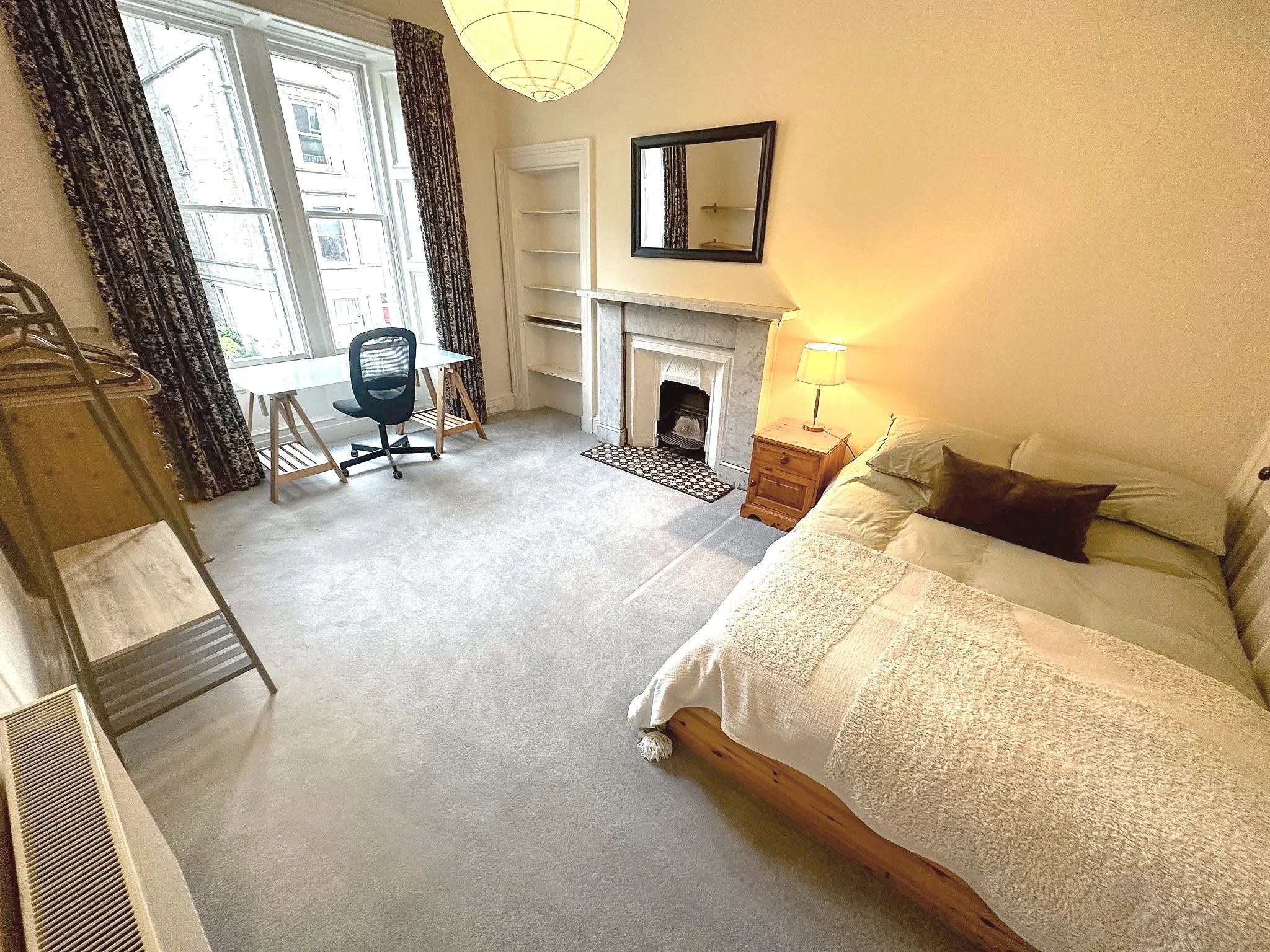 A living room with a bed on the right side, a wooden nightstand and a lamp, a white fireplace with a mirror above, a black chair and a desk near the window, and bookshelves on the left under windows with dark patterned curtains.
