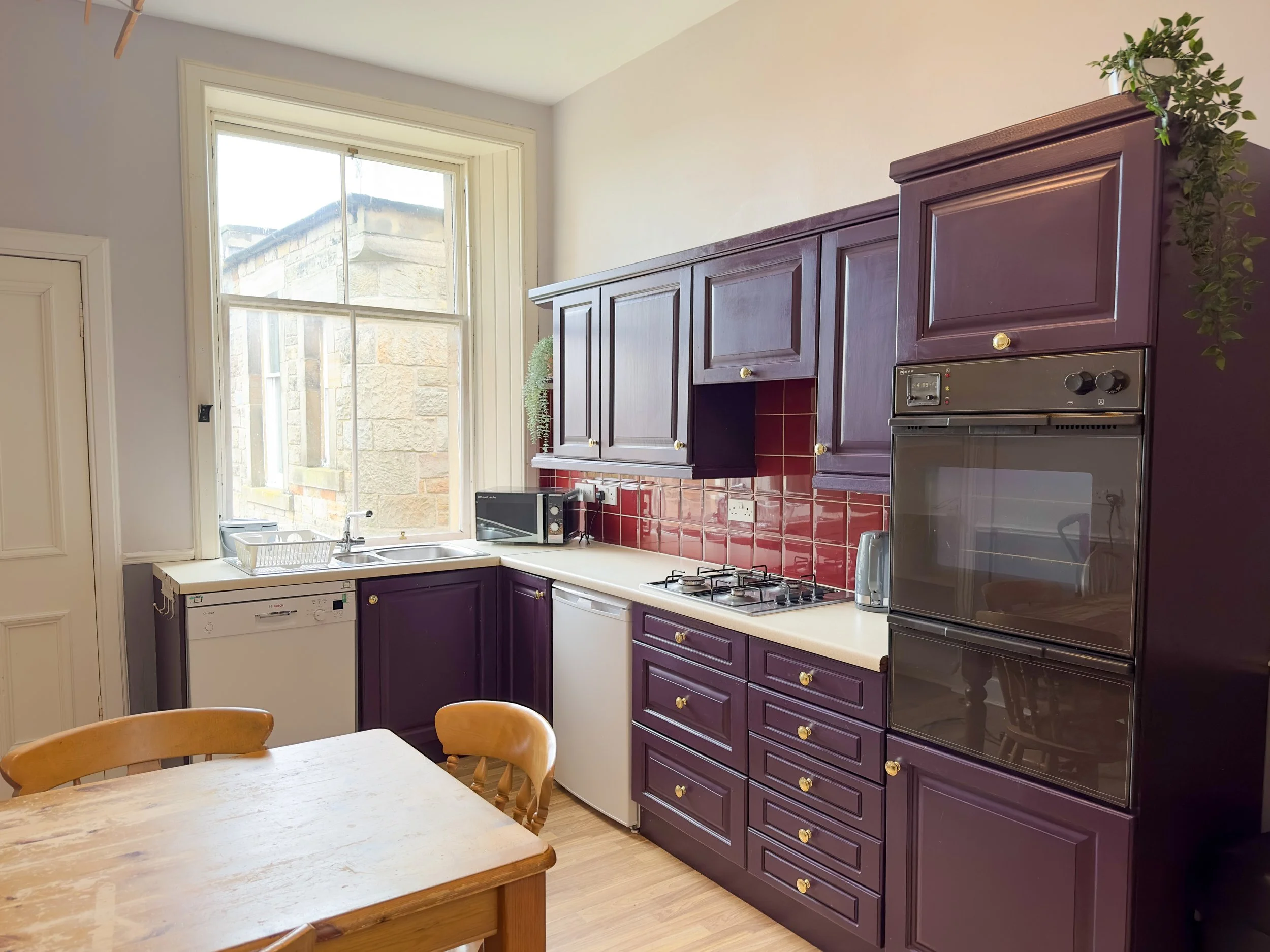 Kitchen with purple cabinets, red tile backsplash, and a large window over the sink. Wooden table and chairs in the foreground.