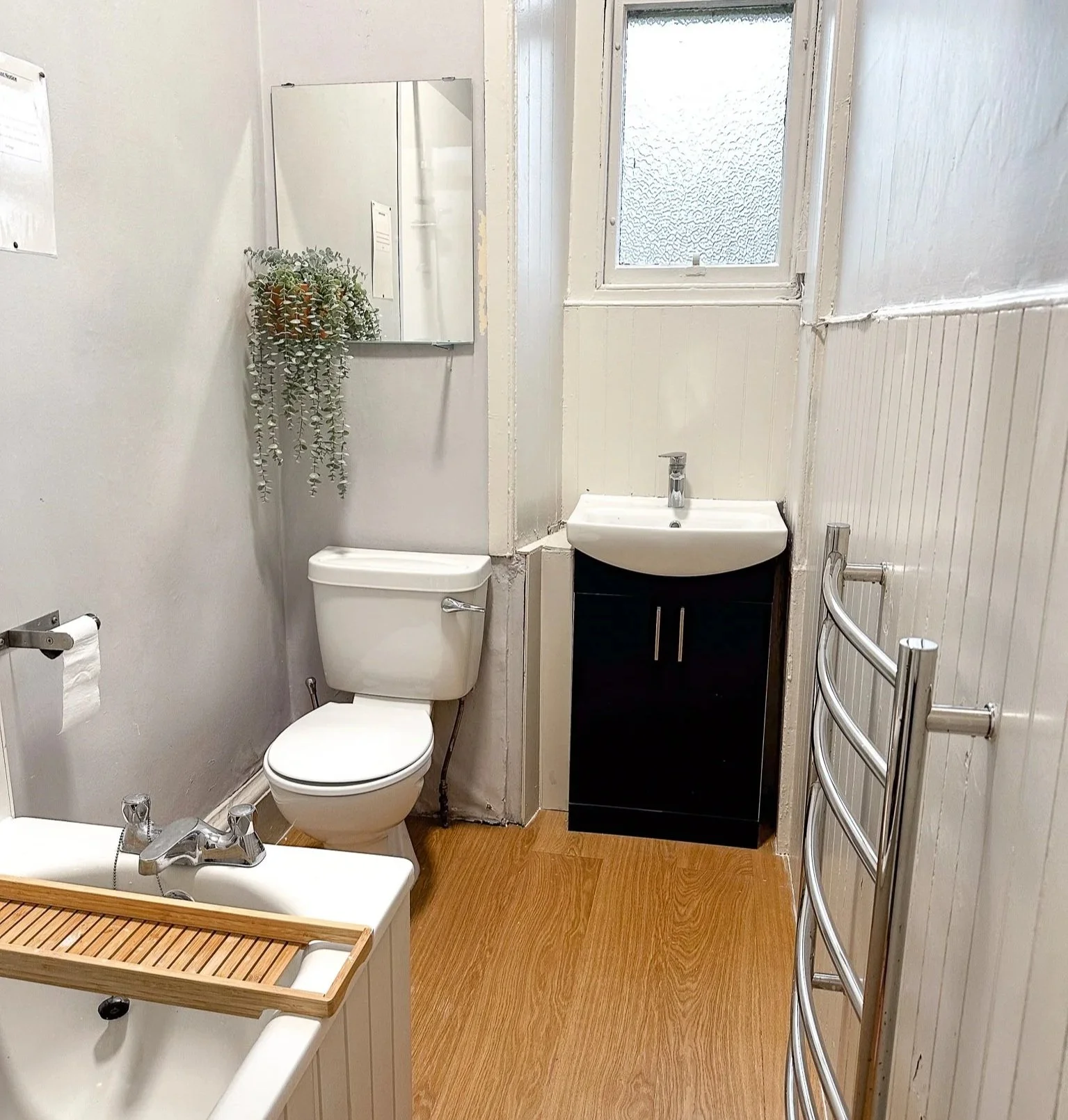 A small bathroom with a white toilet, a black cabinet with a white sink, a small mirror with a hanging plant, a wooden bath tray over a bathtub, a window with textured glass, and a metal towel warmer.