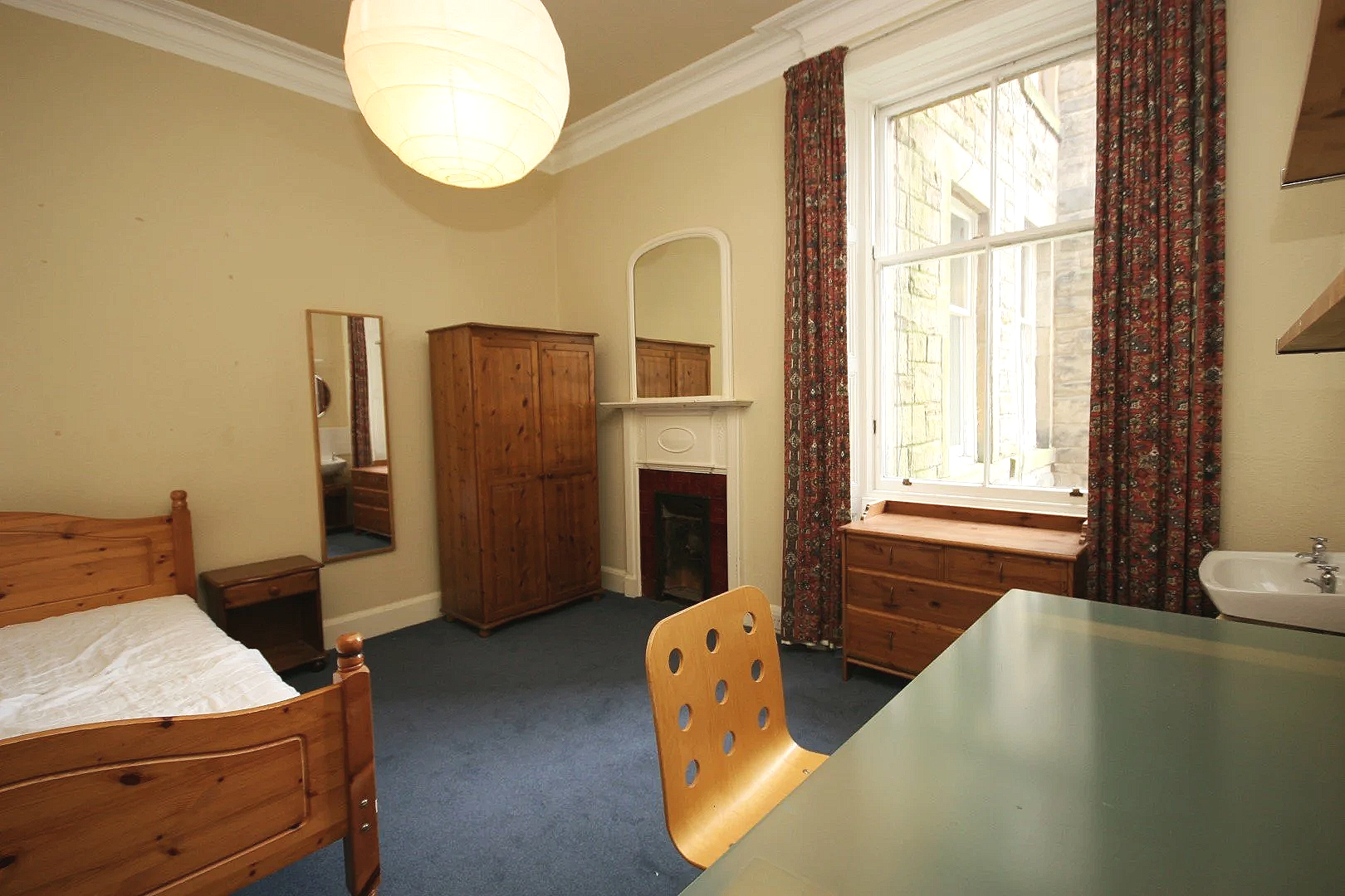 Bedroom with wooden bed and furniture, a window with patterned curtains, a mirror on the wall, a fireplace, and a desk with a chair.