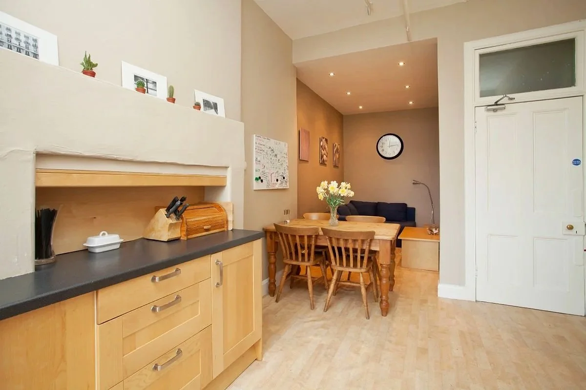 Interior of a kitchen and dining area with light wood cabinets, a black countertop, and a wooden dining table with four chairs. A vase with white flowers sits on the table. In the background, there is a small sitting area with a dark couch, artwork on the wall, a round clock, and a floor lamp. The kitchen has three small potted plants and picture frames on a ledge above the cabinets. The door is white with a small window.