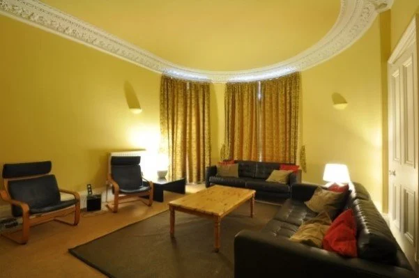 Living room with yellow walls, ornate ceiling, dark leather sofas, wooden chairs, a wooden coffee table, curtains, and lamps.