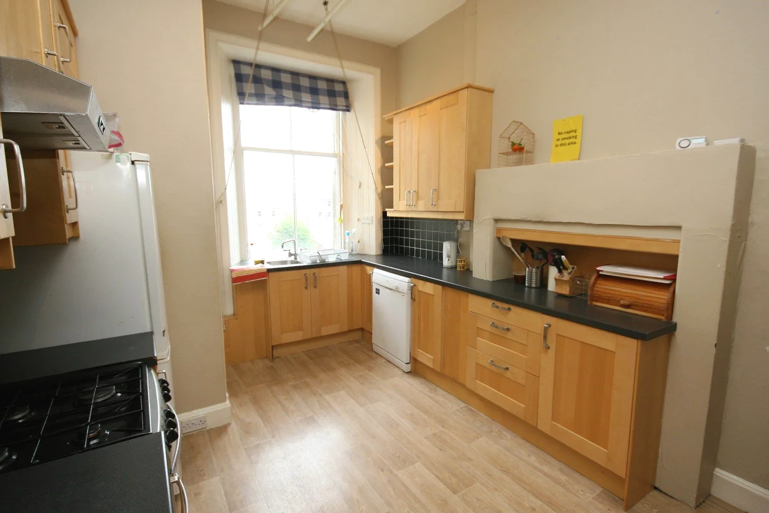 Kitchen with wooden cabinets, black countertop, white dishwasher, stove, window with checkered curtain, and various kitchen items.