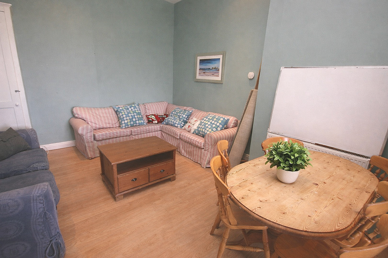 Living room with a pink striped sofa with multiple patterned cushions, a wooden coffee table, a dining table with chairs and a potted plant, and a framed seaside painting on the wall.