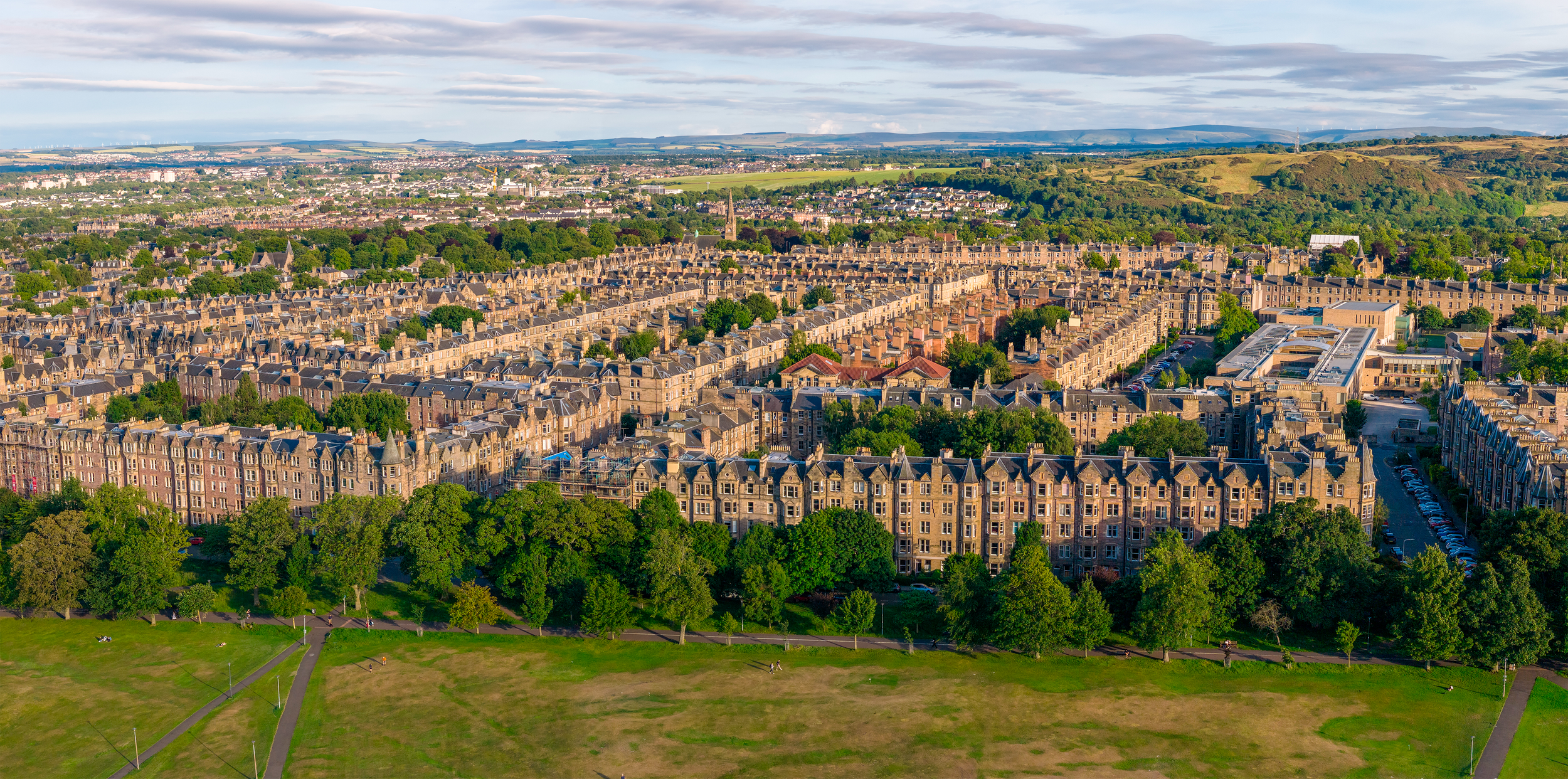 Aerial view of a densely packed residential area with historic buildings, greenery, and a park in the foreground, urban landscape extending to distant hills under a partly cloudy sky.