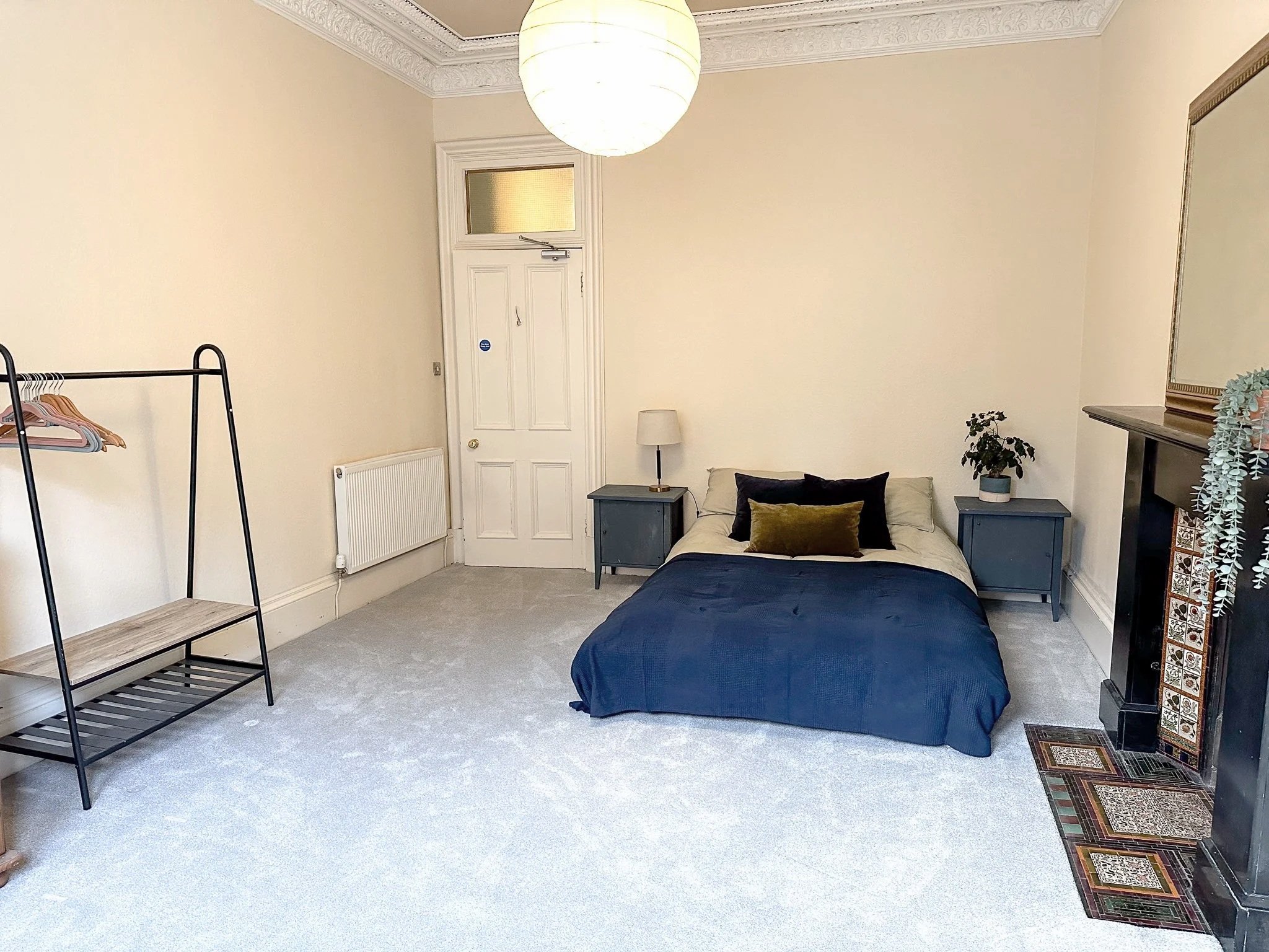 A bedroom with a bed featuring dark blue bedding, alongside two blue nightstands with lamps and a potted plant. There's a white door with a frosted window, a radiator, a black metal clothing rack with hangers, and a fireplace with a decorative tile border on the right side. The ceiling has ornate crown molding and a round paper lantern hanging light.