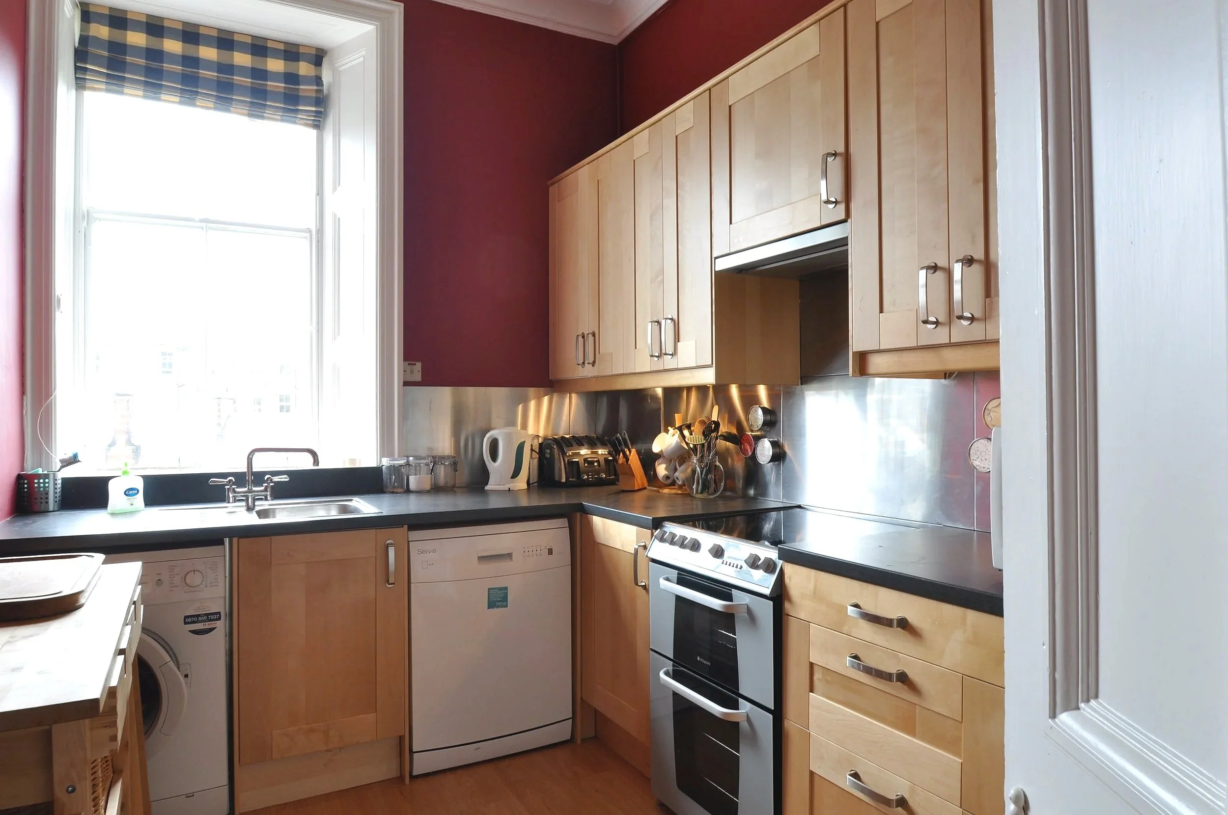 Small kitchen with wood cabinets, black countertops, and a window with a plaid valance. Contains a white washing machine, a dishwasher, an electric stove, a kettle, and various kitchen utensils and containers.