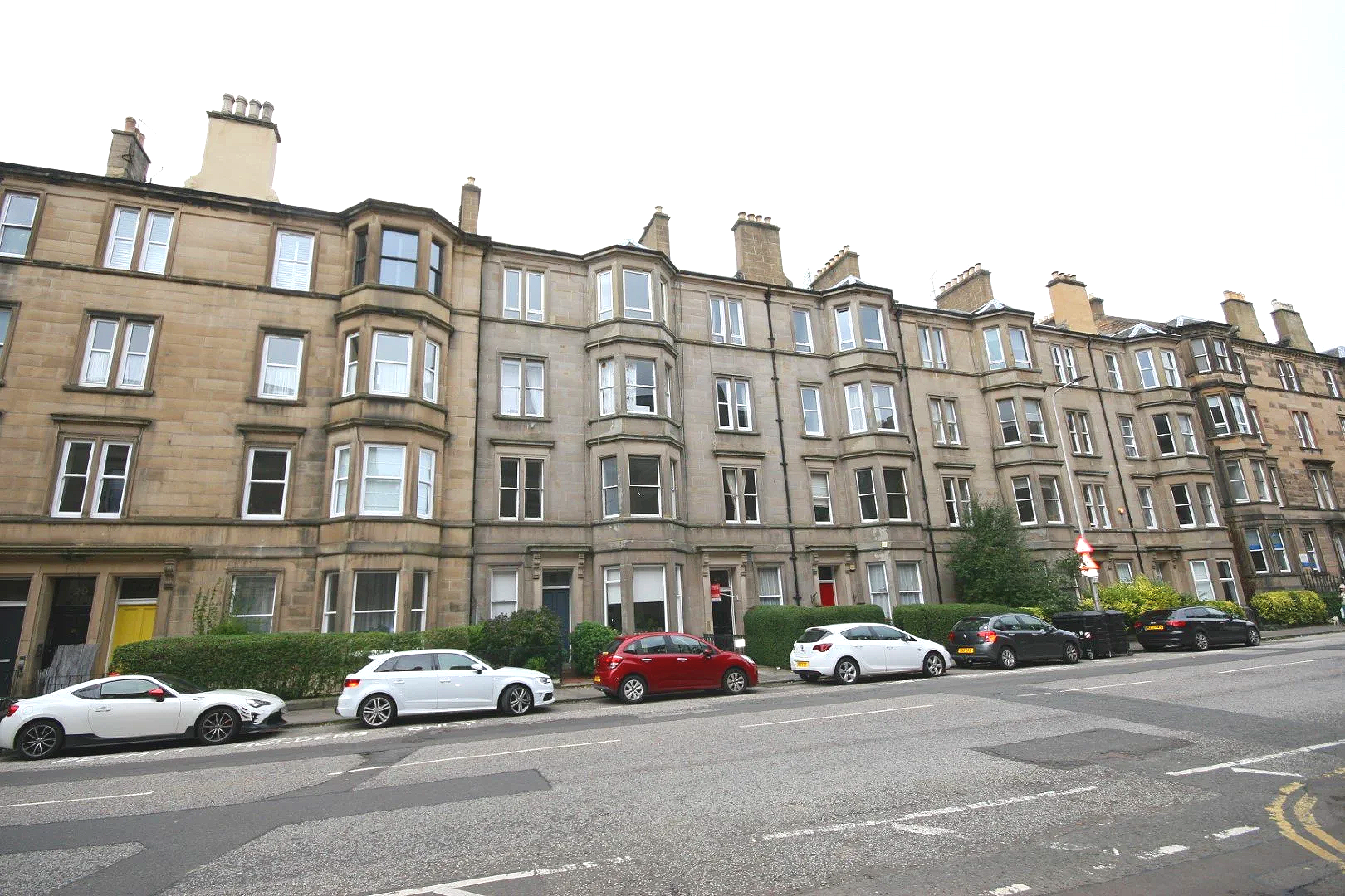 A corner view of a sandstone multi-story apartment building with bay windows, on a city street with parked cars and a blue sky with clouds.