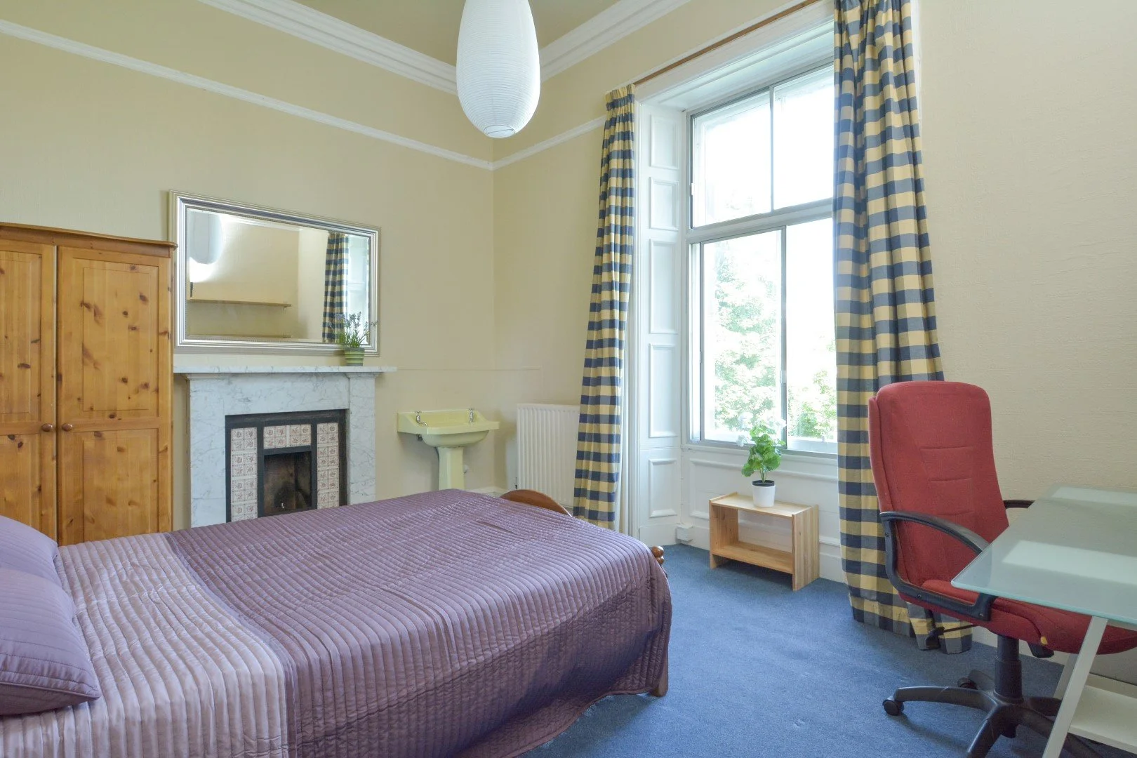 Bright bedroom with a bed, upright red office chair, large window with plaid curtains, small wooden table with a plant, fireplace with a mirror above, and a wooden wardrobe.