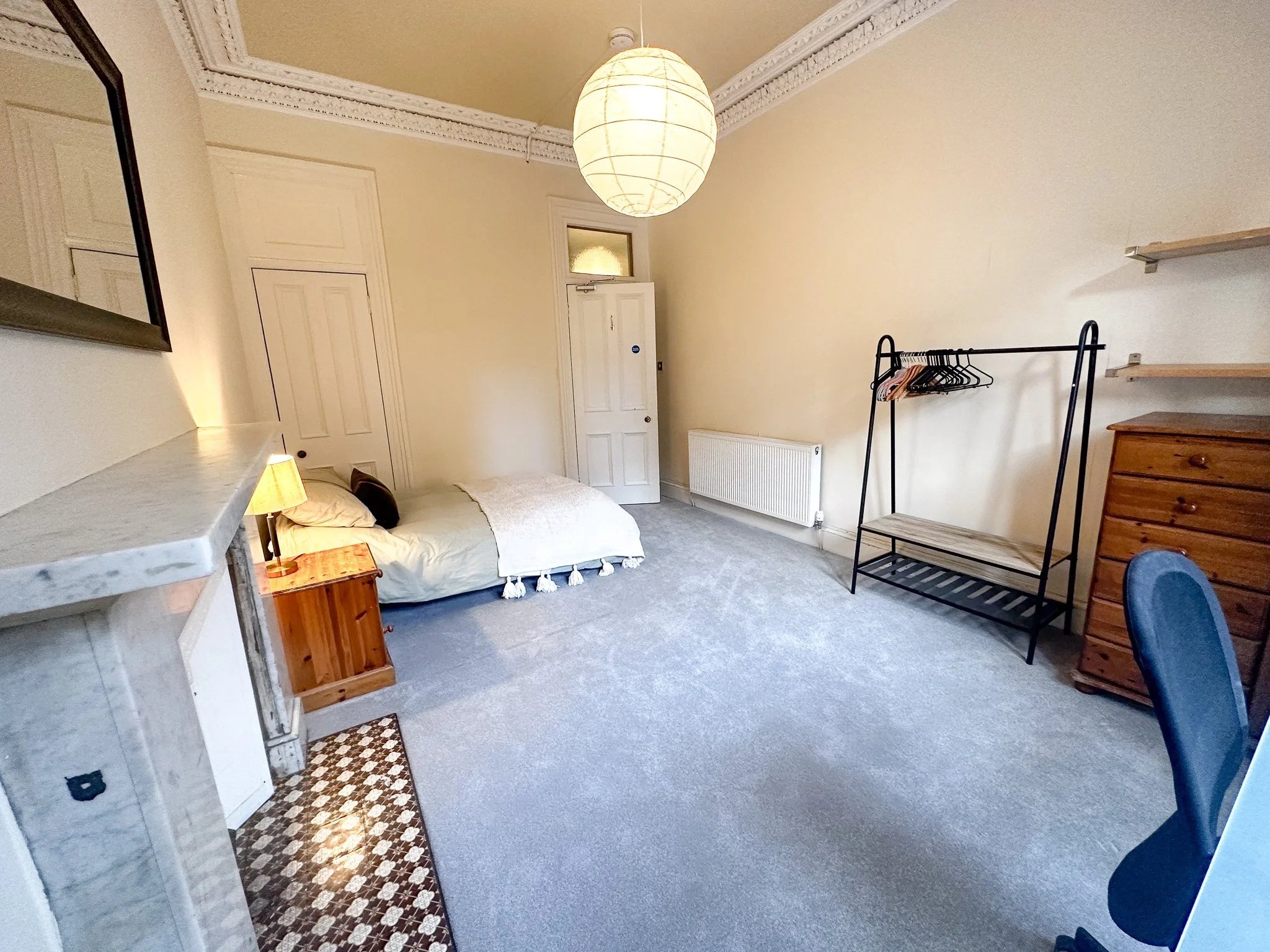 A spacious bedroom with a bed on the left side near the wall, a small wooden nightstand with a lamp, built-in white closet doors, beige walls, a patterned rug near the fireplace, a coat rack with hangers, a wooden dresser, open wall shelves, a gray carpeted floor, a radiator, and a paper lantern ceiling light.