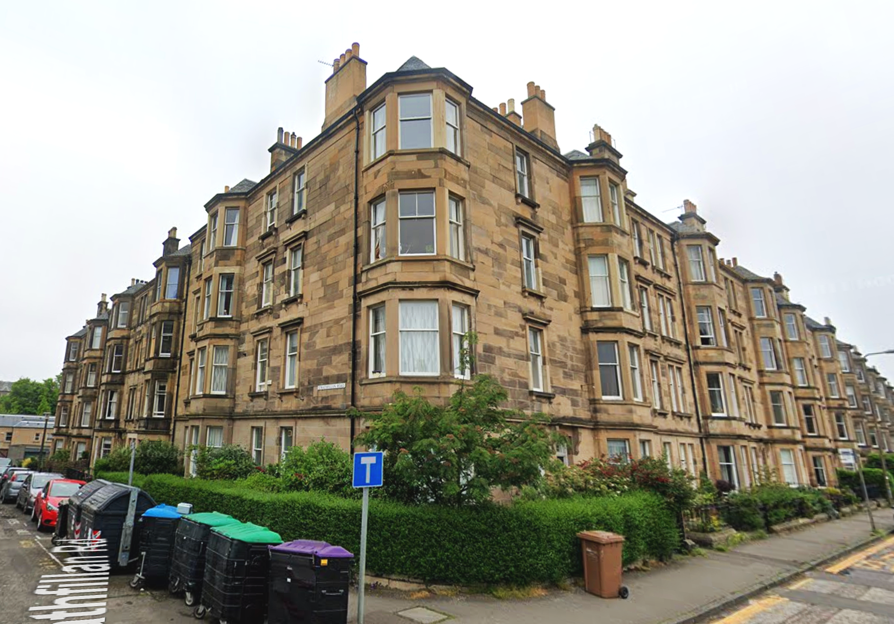 A multi-story sandstone residential building on a street corner, with bay windows, small gardens, and parked cars in the foreground, under an overcast sky.