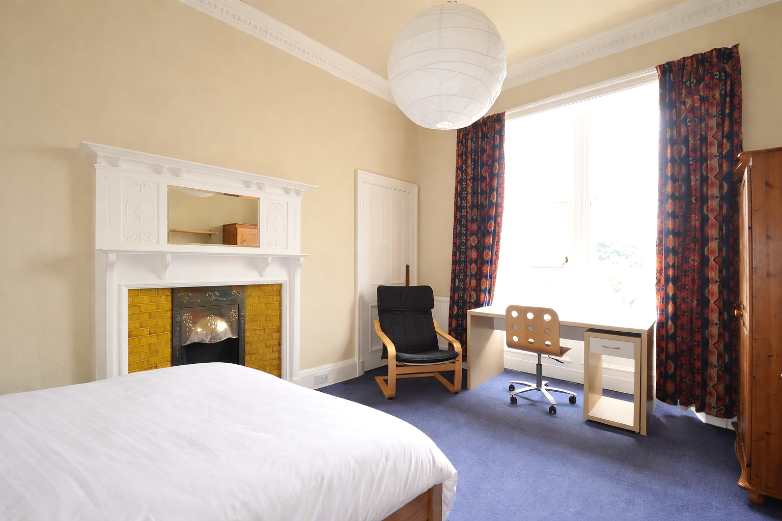 A bedroom with a white bed, a fireplace with a mirror, a black armchair, a white desk, a swivel chair, dark patterned curtains, and a wooden wardrobe.