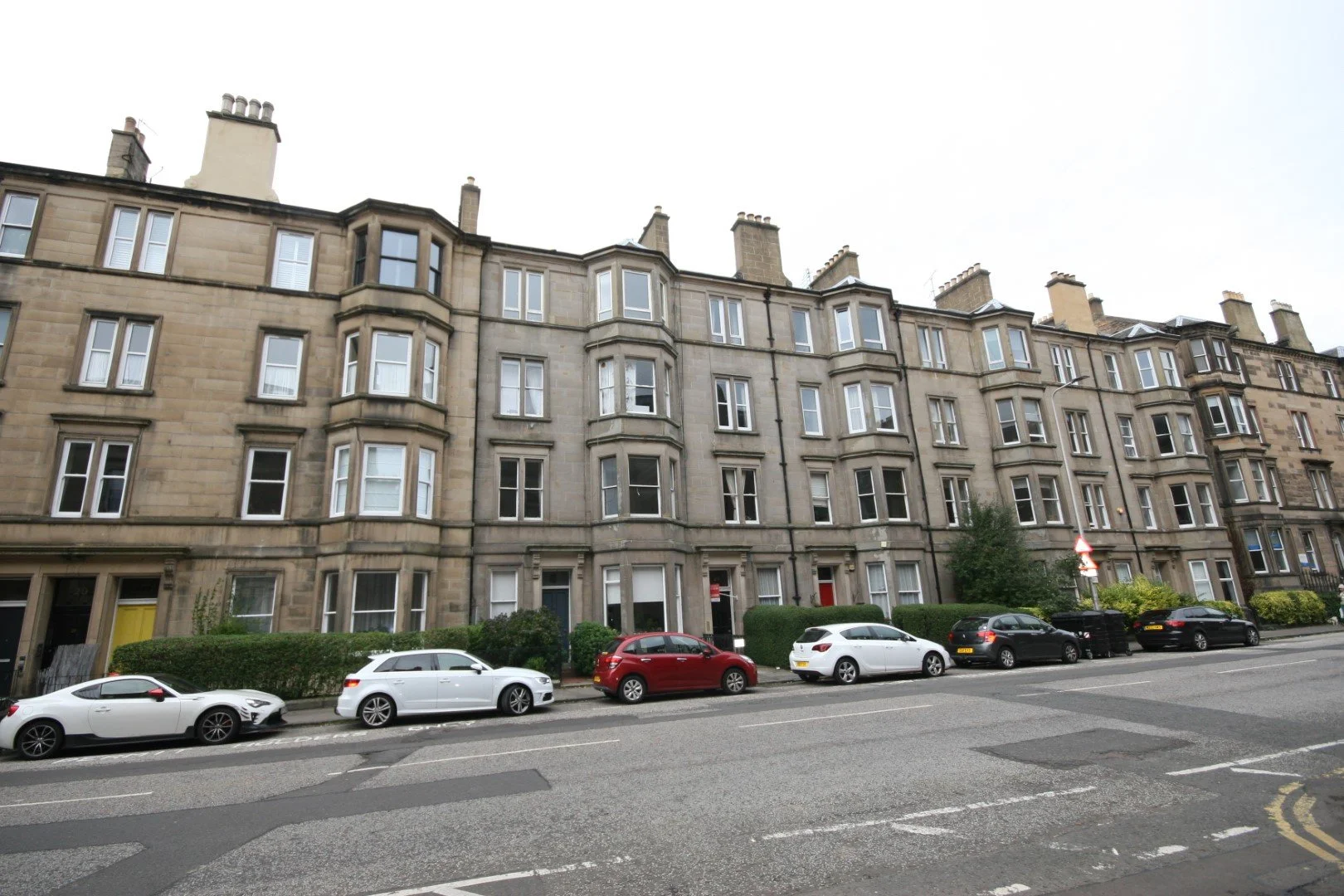 A corner view of a sandstone multi-story apartment building with bay windows, on a city street with parked cars and a blue sky with clouds.